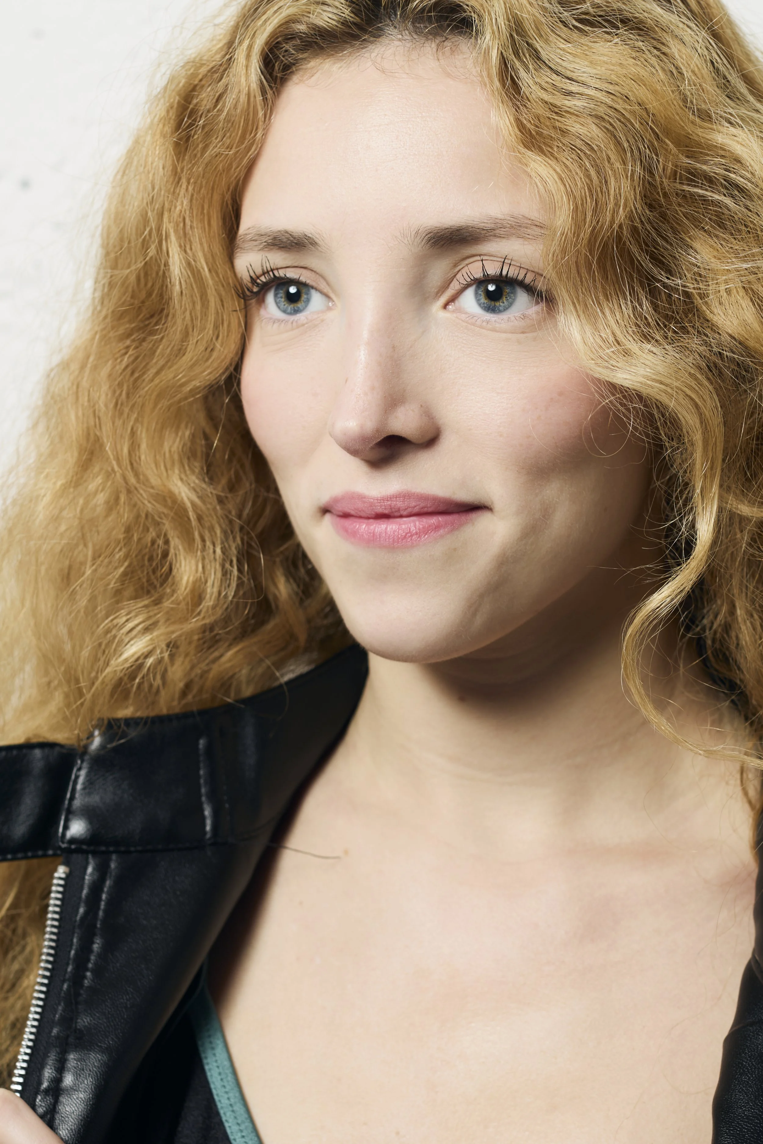 Close-up of a woman with long, curly blonde hair, blue eyes, and light skin, wearing a black leather jacket.