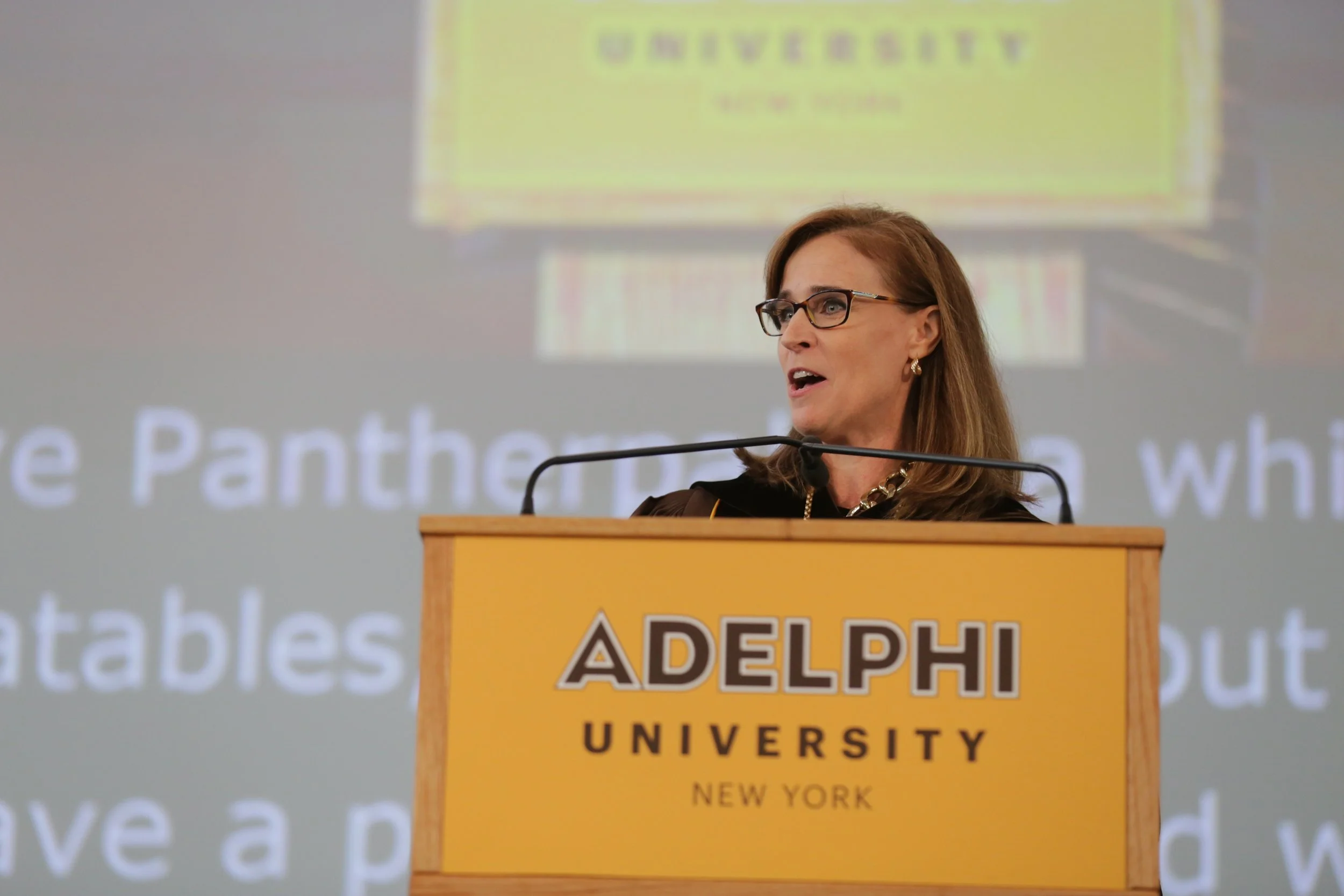 A woman with brown hair, glasses, and wearing earrings and a black top stands at a podium with a yellow sign reading 'ADELPHI UNIVERSITY NEW YORK.' She is speaking at an event, with a blurred background showing a projection featuring the word 'GIVENE