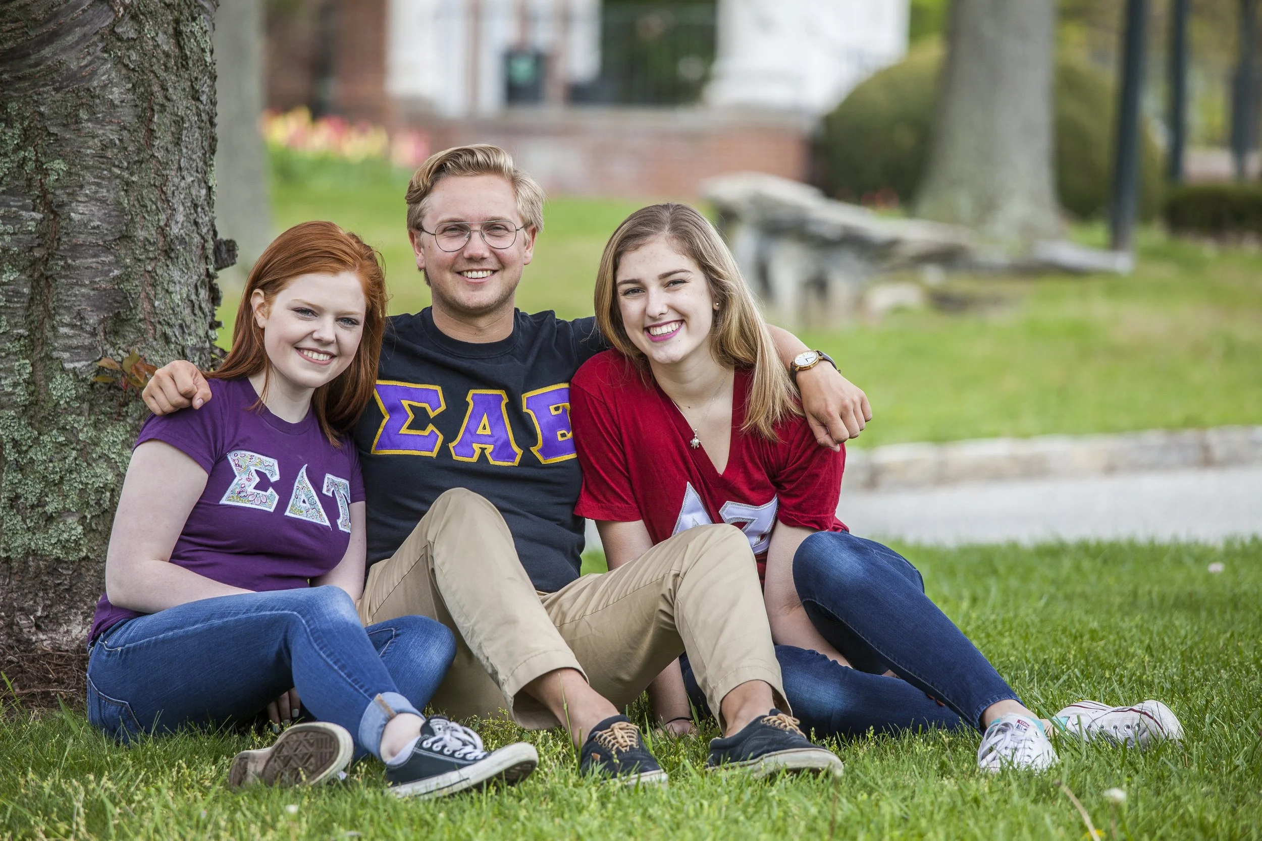 Three smiling friends, two women and one man, sitting under a tree on grass. The women are wearing sorority shirts, and the man is wearing a Sigma Alpha Epsilon shirt. They are outdoors in a park-like setting.