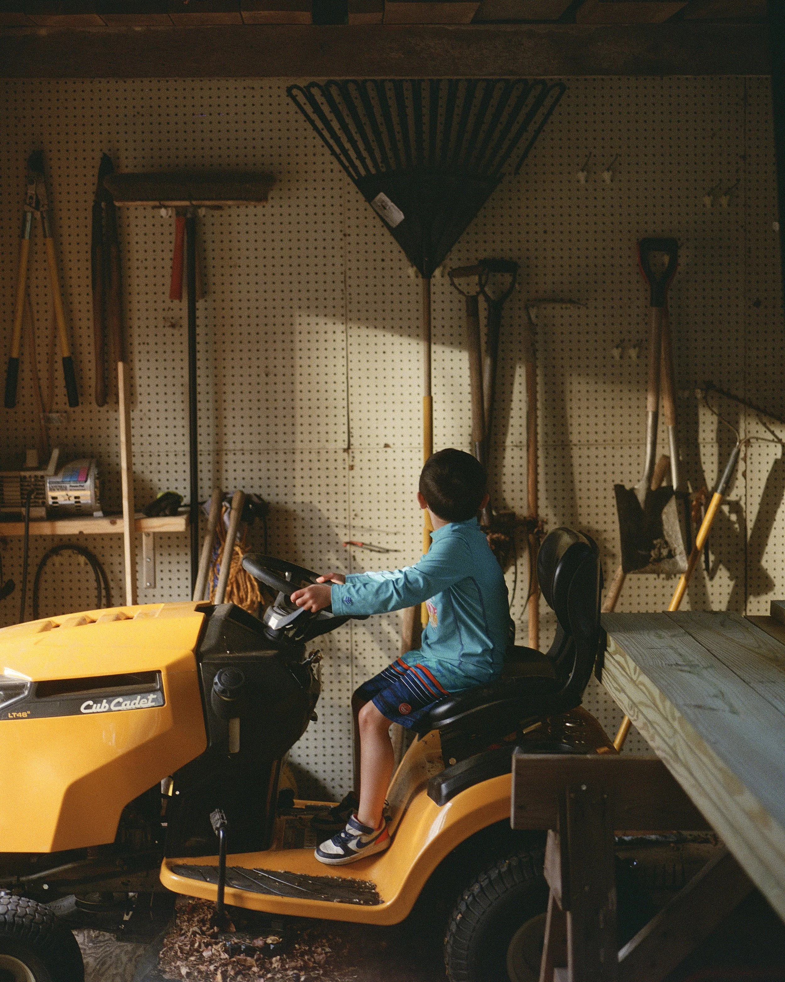 A young boy wearing a turquoise jacket, plaid shorts, and sneakers is seated on a yellow Cub Cadet lawn tractor inside a garage. The garage has a pegboard wall with various garden tools hanging, including a rake, shovels, and a broom. The scene featu