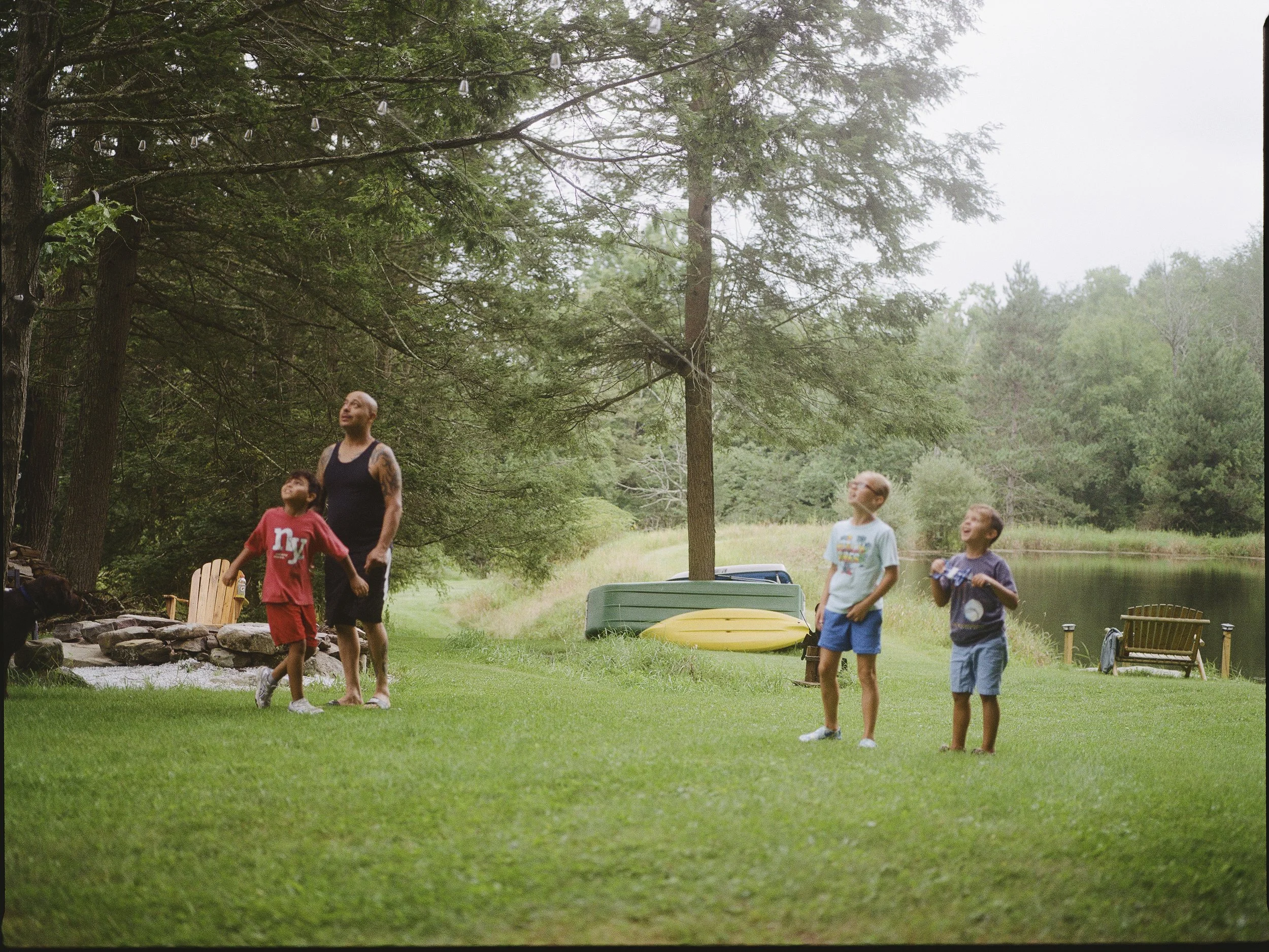 A group of children and a man outdoors in a lush, green park with tall trees. The children are playing, and the man is standing nearby, watching them.