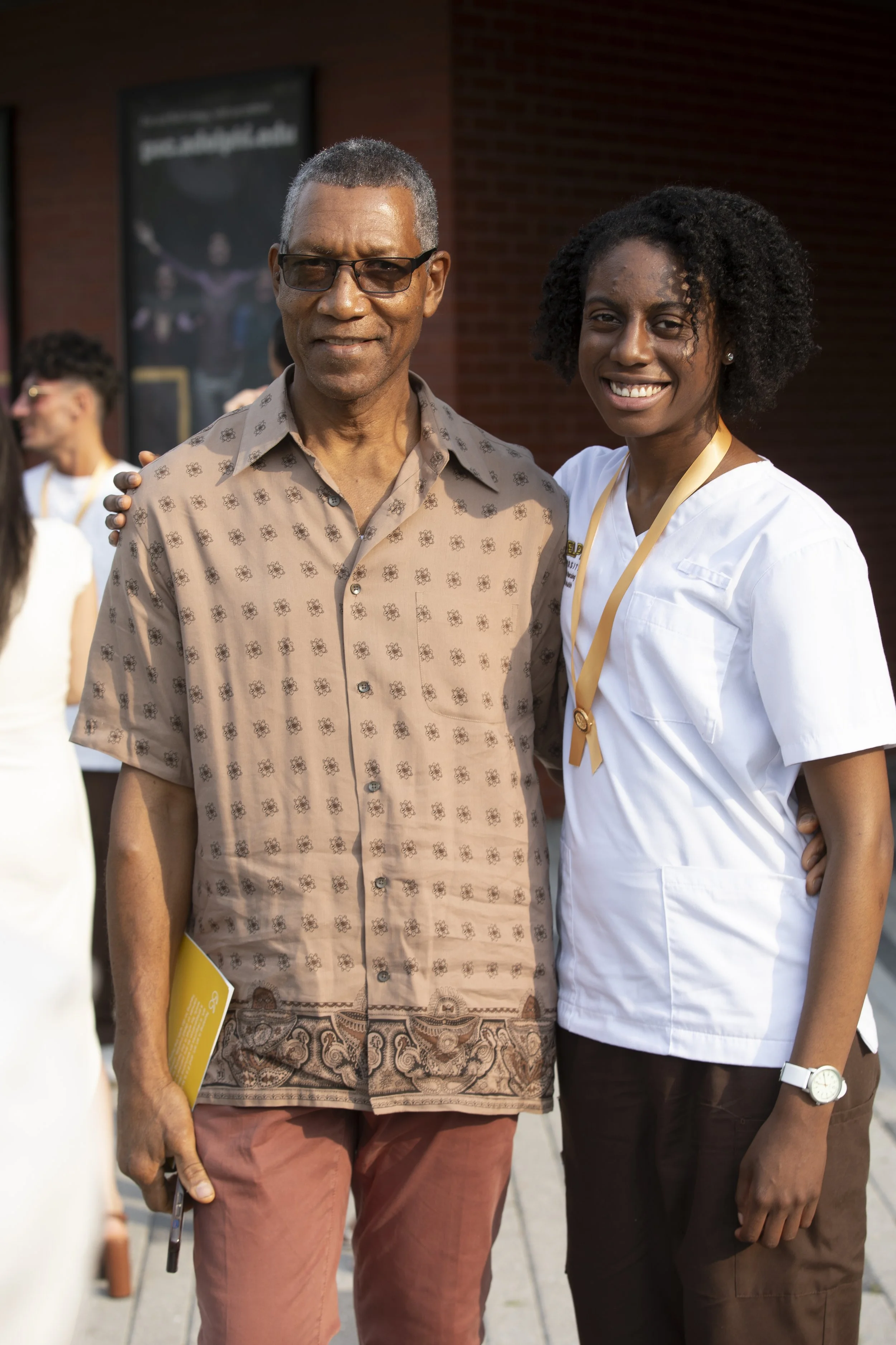 Smiling man and woman in white medical uniform posing outdoors at a public event, with other people in the background.