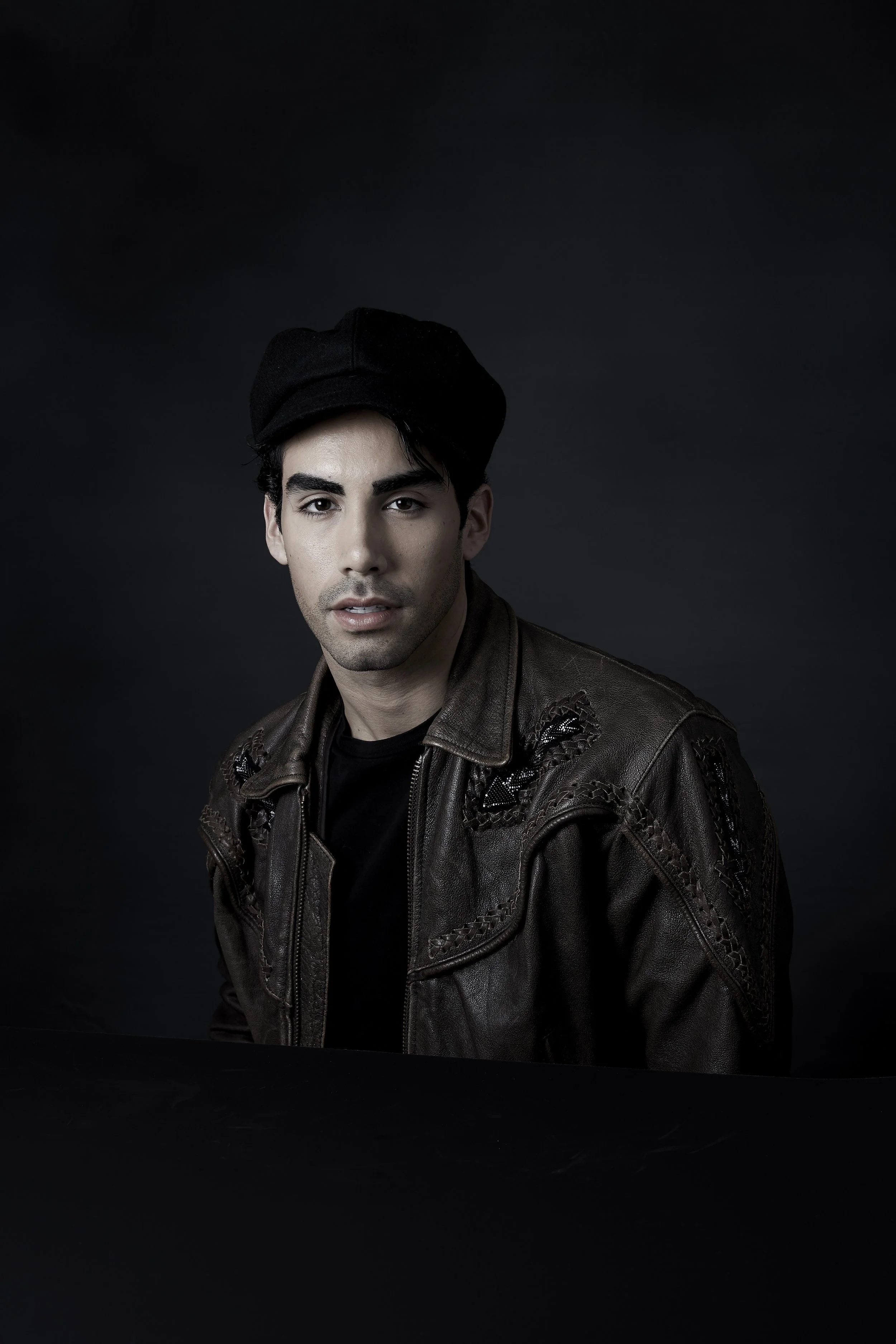 Portrait of a man wearing a black hat and a brown leather jacket against a dark background.