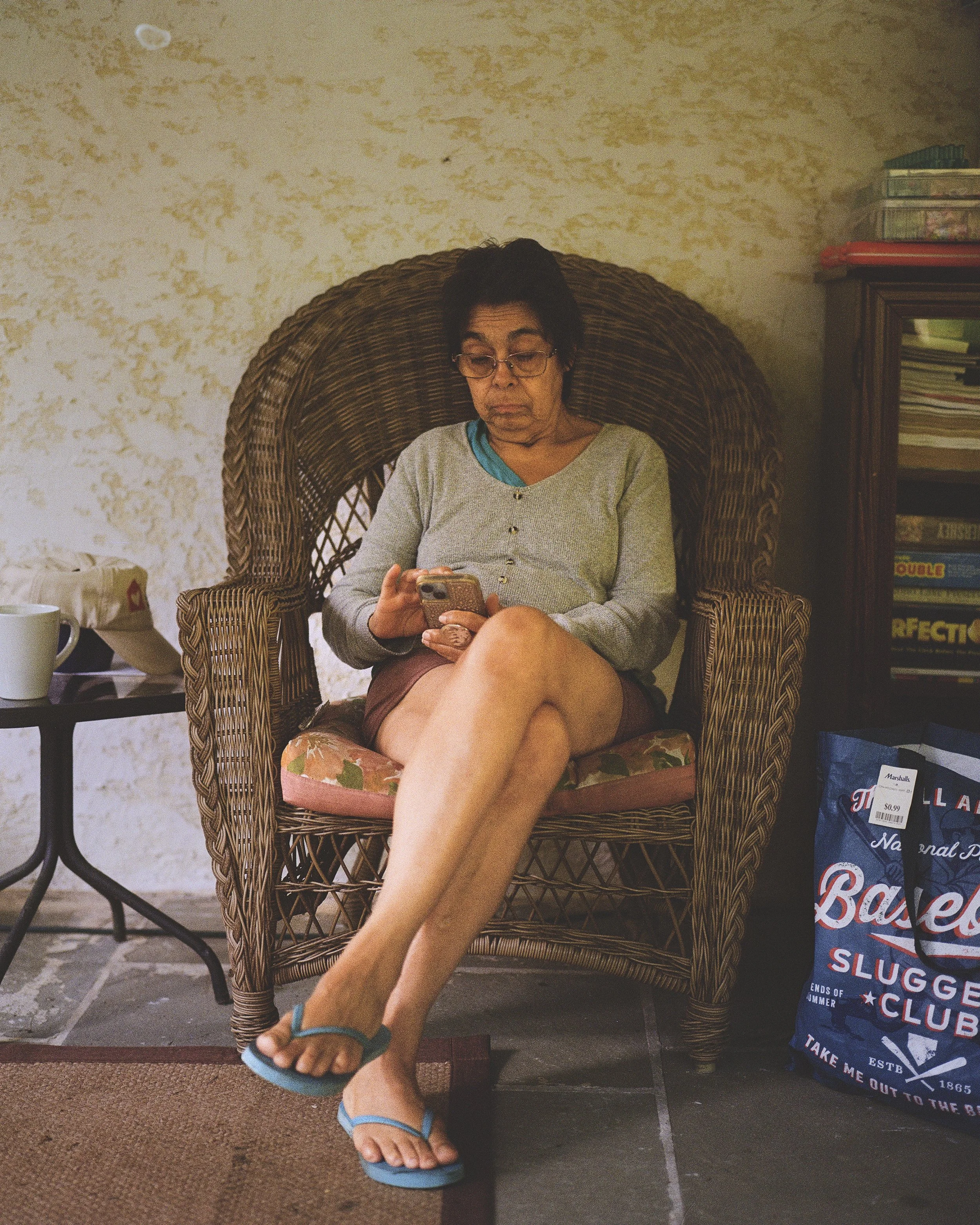 A woman with short dark hair and glasses sitting in a wicker chair, looking at her phone. She is wearing a light gray cardigan, a blue shirt, and sandals. The setting appears to be indoors or on a patio, with a small table to her right holding a whit