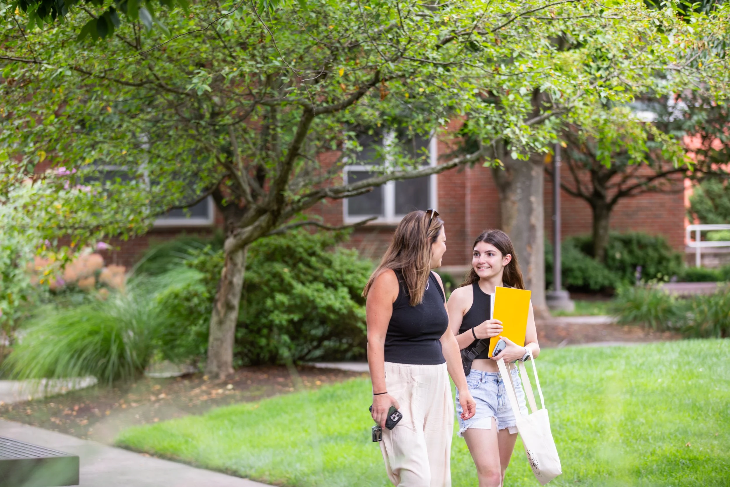 Two young women walking and talking in a park, with trees and a brick building in the background. One is holding a phone, the other a yellow folder and a tote bag.