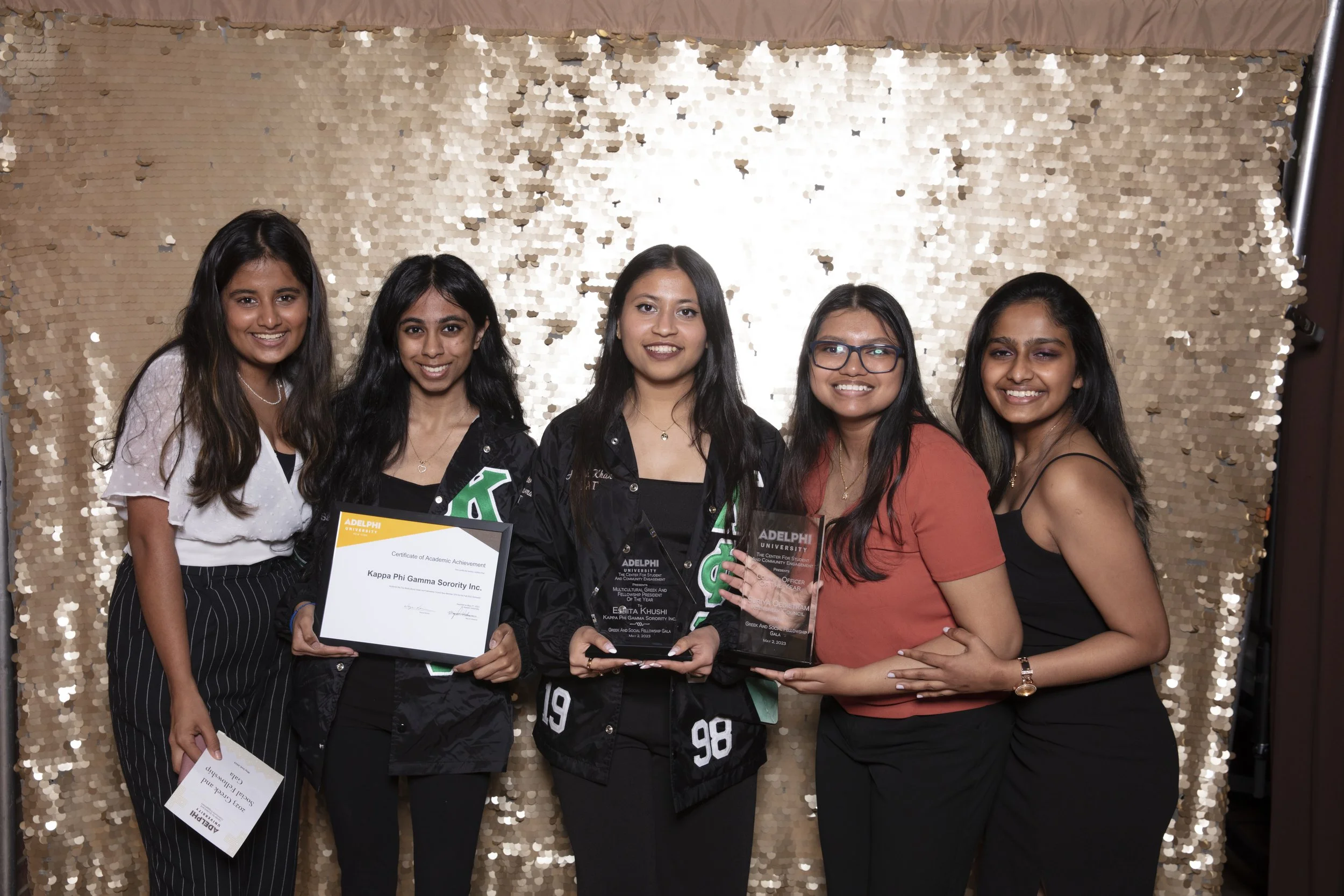 Five young women standing together, smiling, with some holding awards and certificates, in front of a gold sequin backdrop.