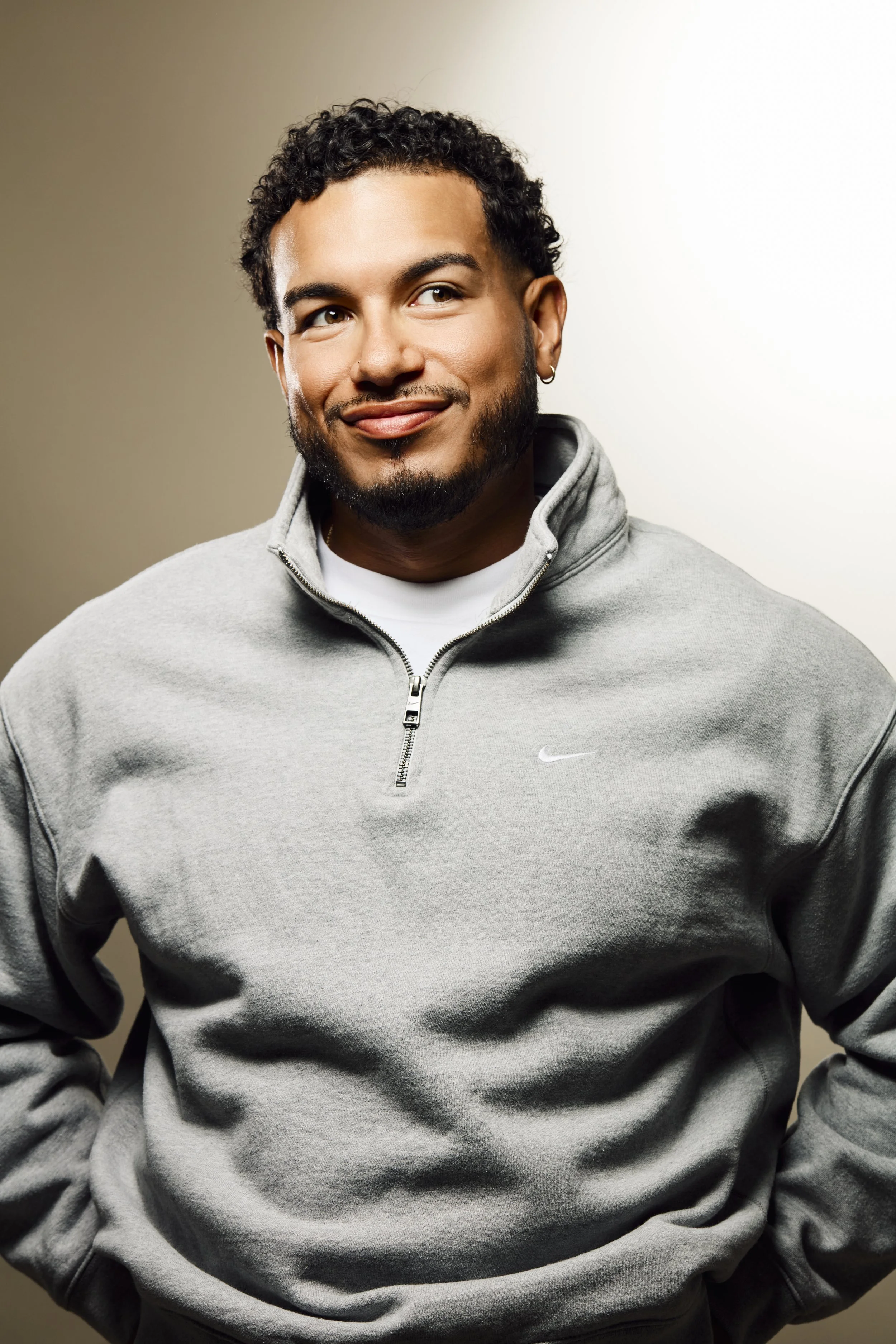 A young man with curly hair and a beard, wearing a grey Nike hoodie, smiling and looking to the side, against a neutral background.
