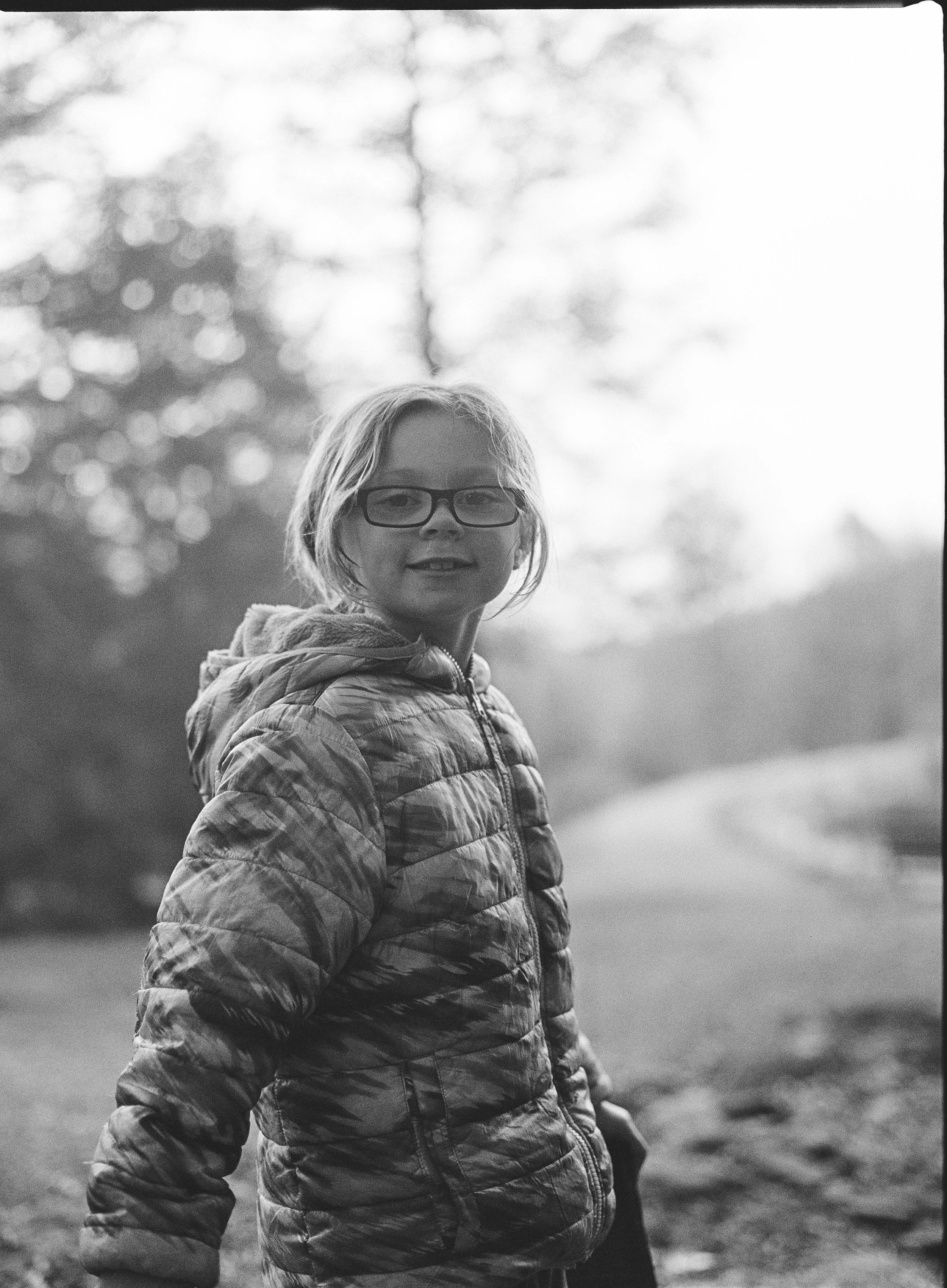 A smiling girl with glasses and a puffy jacket outdoors, with trees and a cloudy sky in the background.