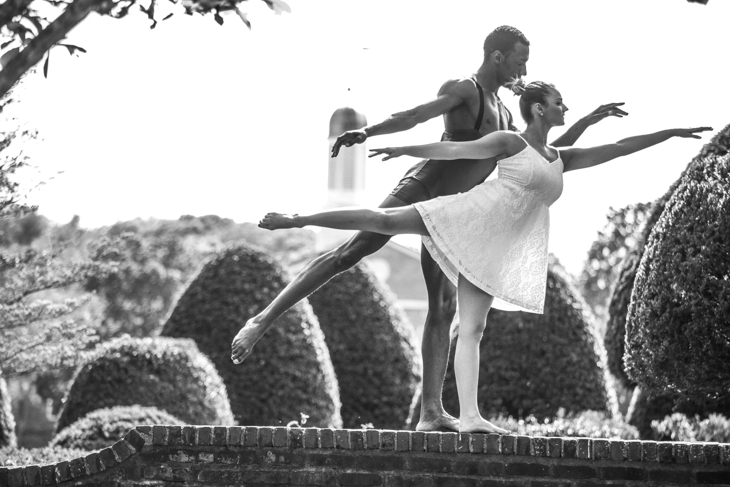 Two ballet dancers practicing outdoors, balancing on one leg with arms extended, on a brick ledge in front of shaped bushes, with a tall building in the background.