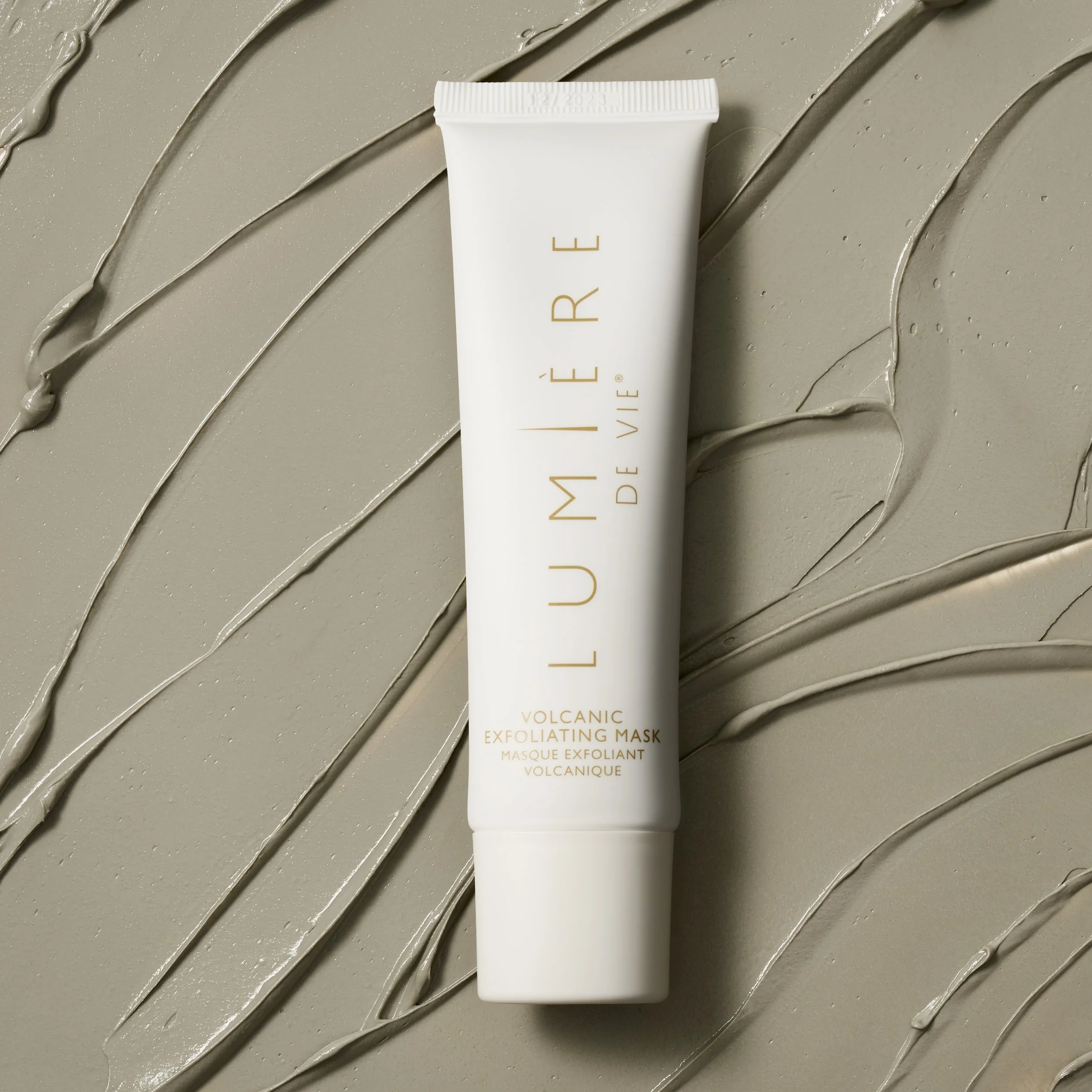 A white tube of LUMIÈRE De VIE volcanic exfoliating mask on a beige surface with texture and streaks of the mask spread across.