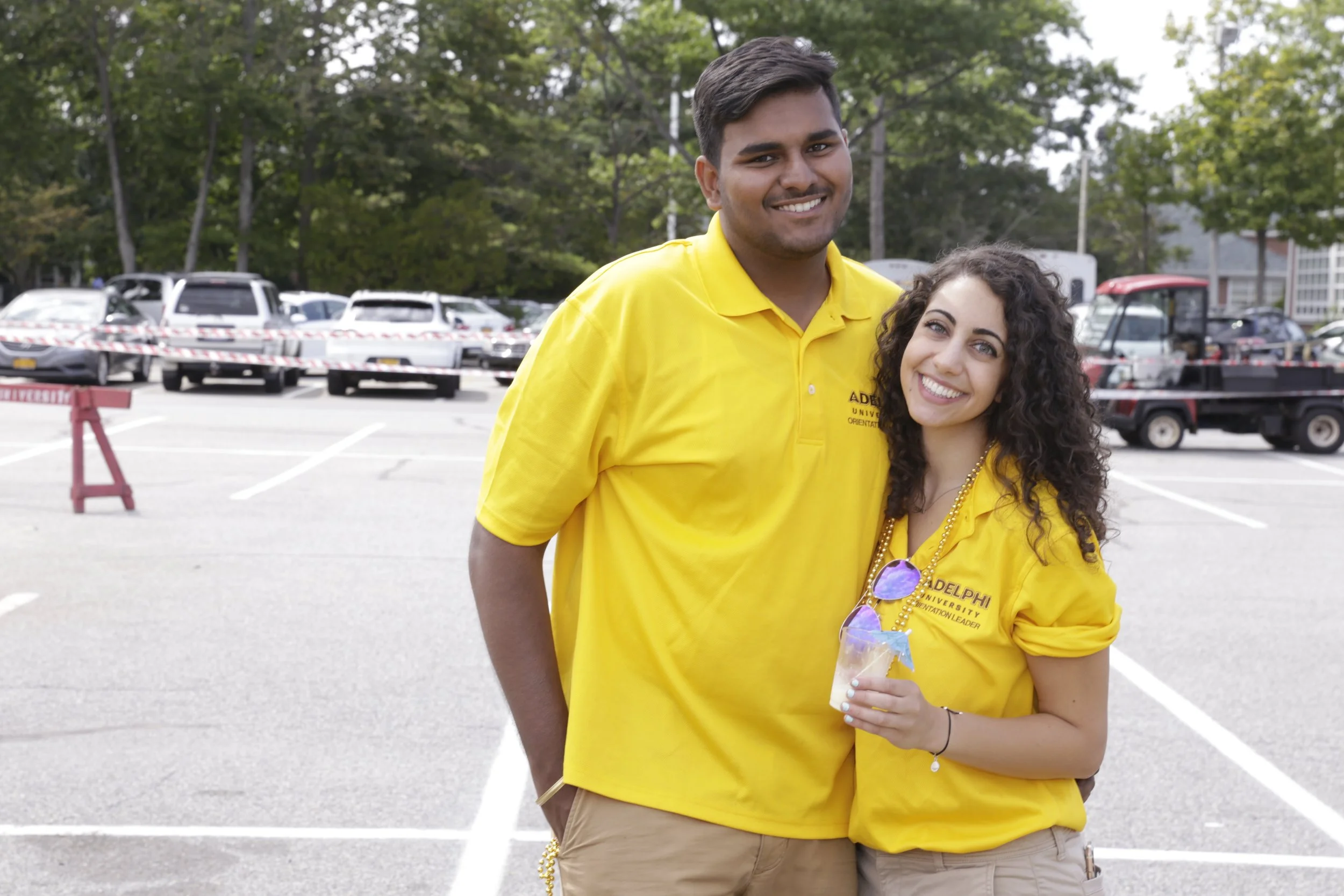 Two young people standing in a parking lot, smiling at the camera, wearing yellow polo shirts from a university, with cars and trees in the background.