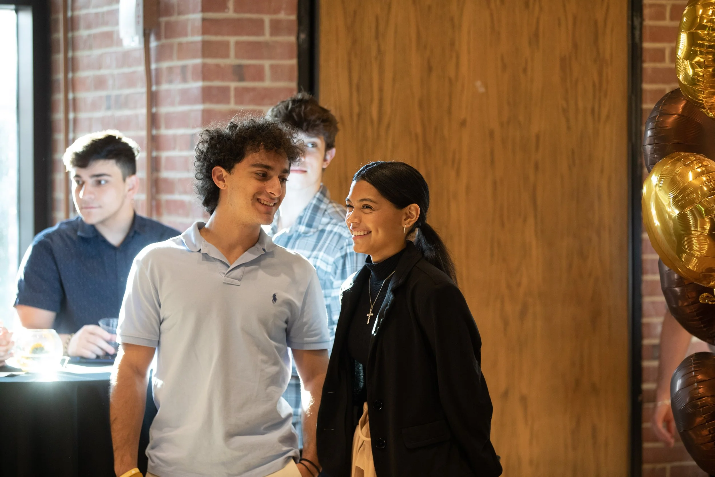 Two young adults, a man and a woman, smiling at each other at a social gathering, with two other young men in the background and decorations including gold balloons.