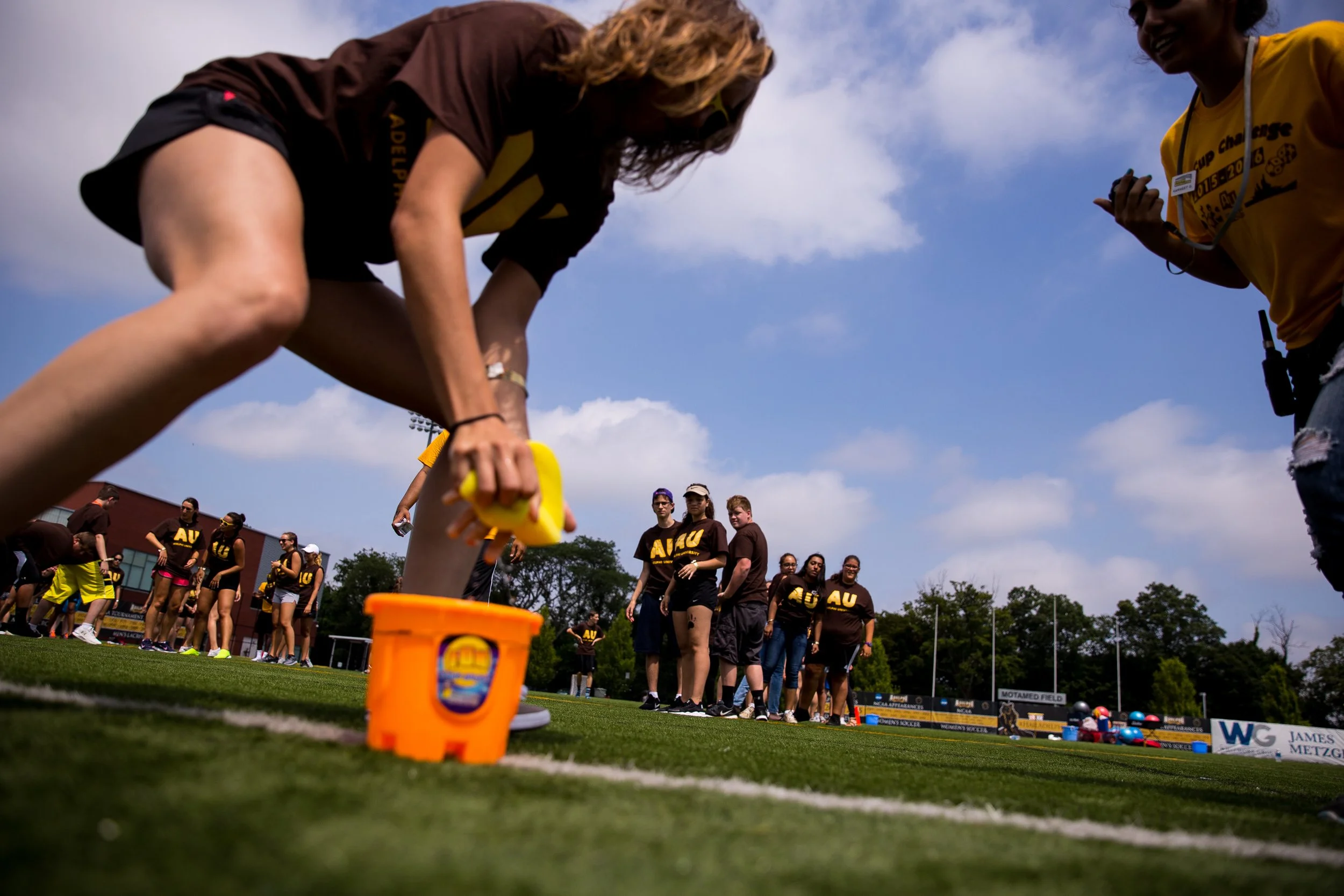 Group of people gathered outdoors on a sports field, participating in an event involving small colored helmets and a yellow bucket, with a woman in the foreground reaching into the bucket, and others watching or waiting in line.