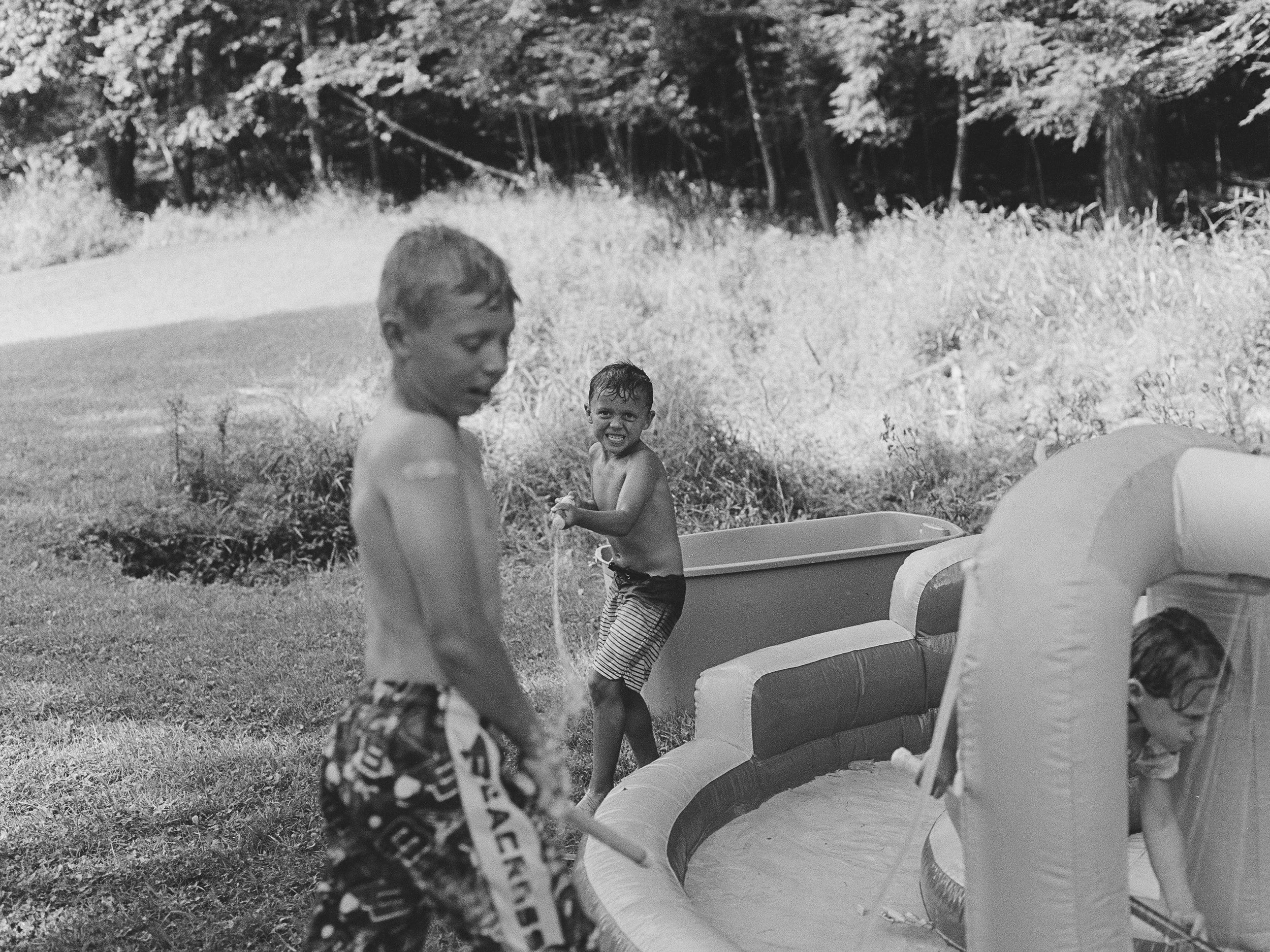 Two children playing outdoors near a large inflatable pool, surrounded by grass and trees in the background. One boy is smiling and wearing striped shorts, while the other is standing shirtless with patterned shorts.
