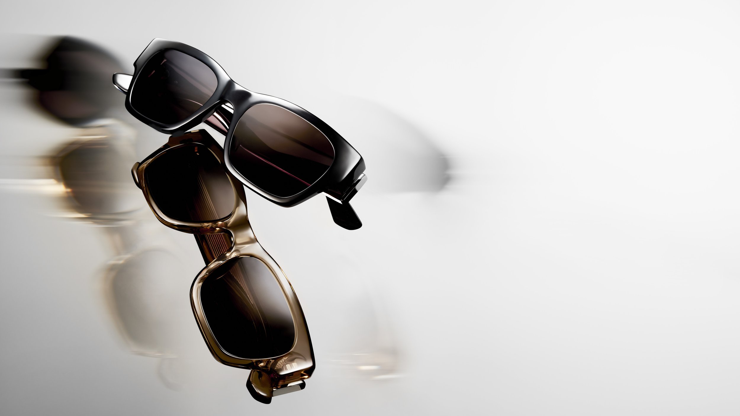 Two pairs of sunglasses, one black and one brown, are stacked and reflect light, placed on a white surface, with shadows cast underneath.