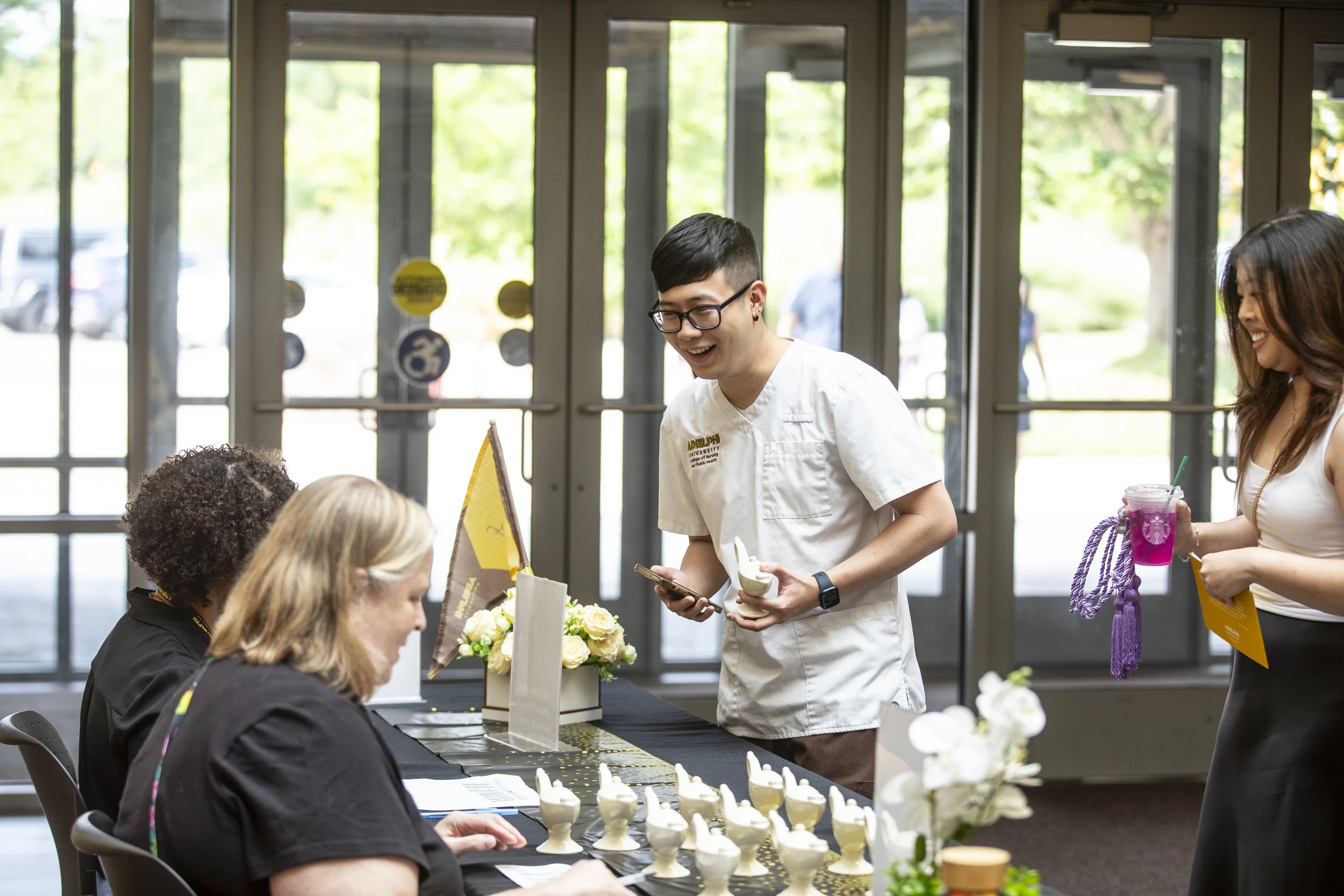 Group of people at a registration table during an event, with a young man in a white shirt smiling and talking, holding a phone and a dessert, while two women sit at the table and another woman stands holding a colorful drink.