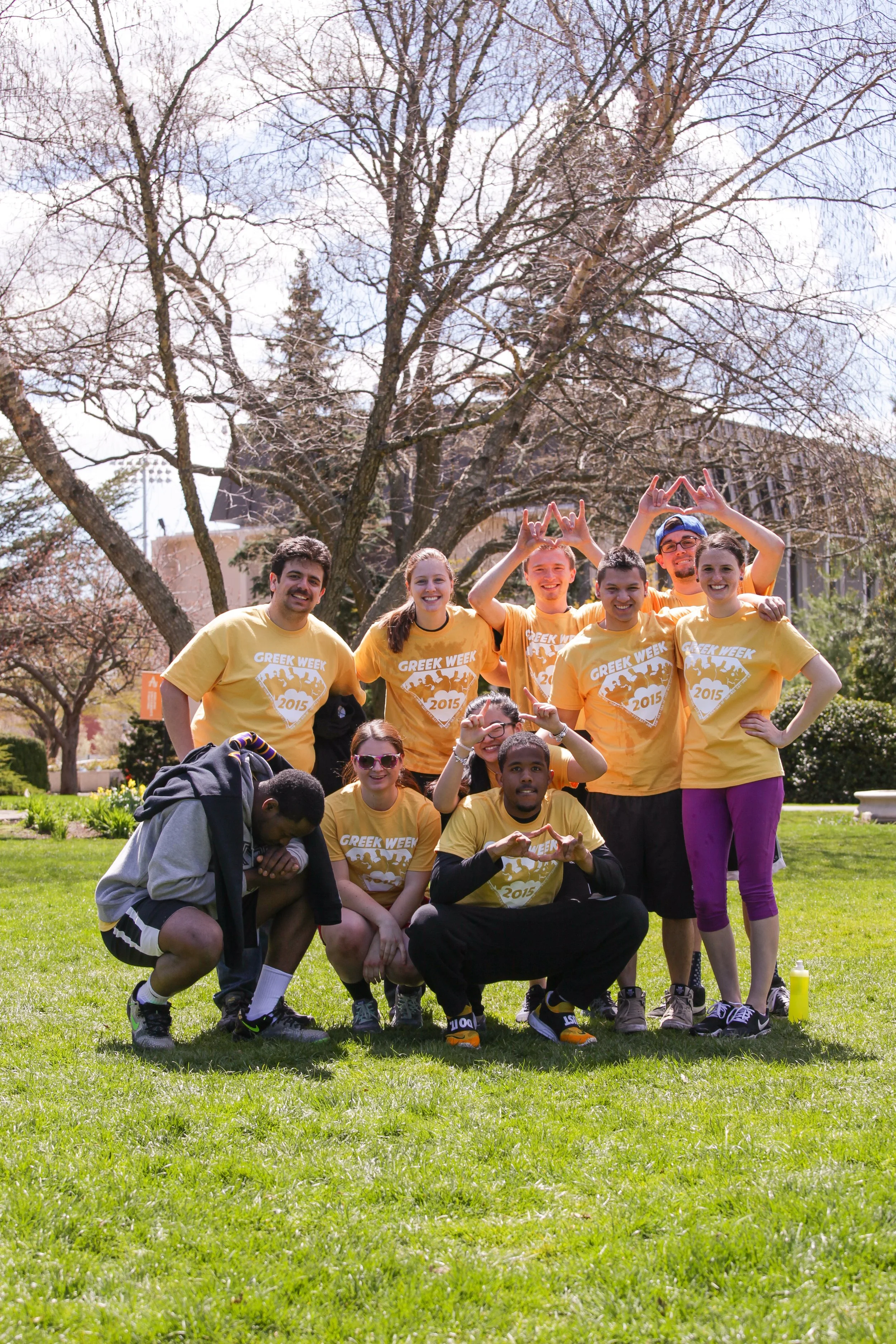 A group of ten people wearing yellow T-shirts posing outdoors on a green lawn during a sunny day. The T-shirts read "Greek Week 2015." They are smiling, making heart signs and other hand gestures, with some individuals crouching and others standing b