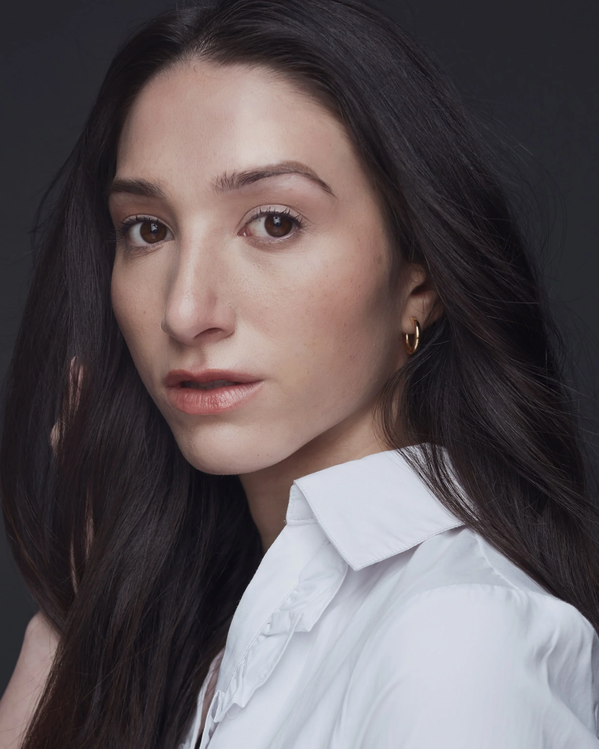 A woman with long dark hair, wearing a white shirt and gold earrings, is posing against a dark background.