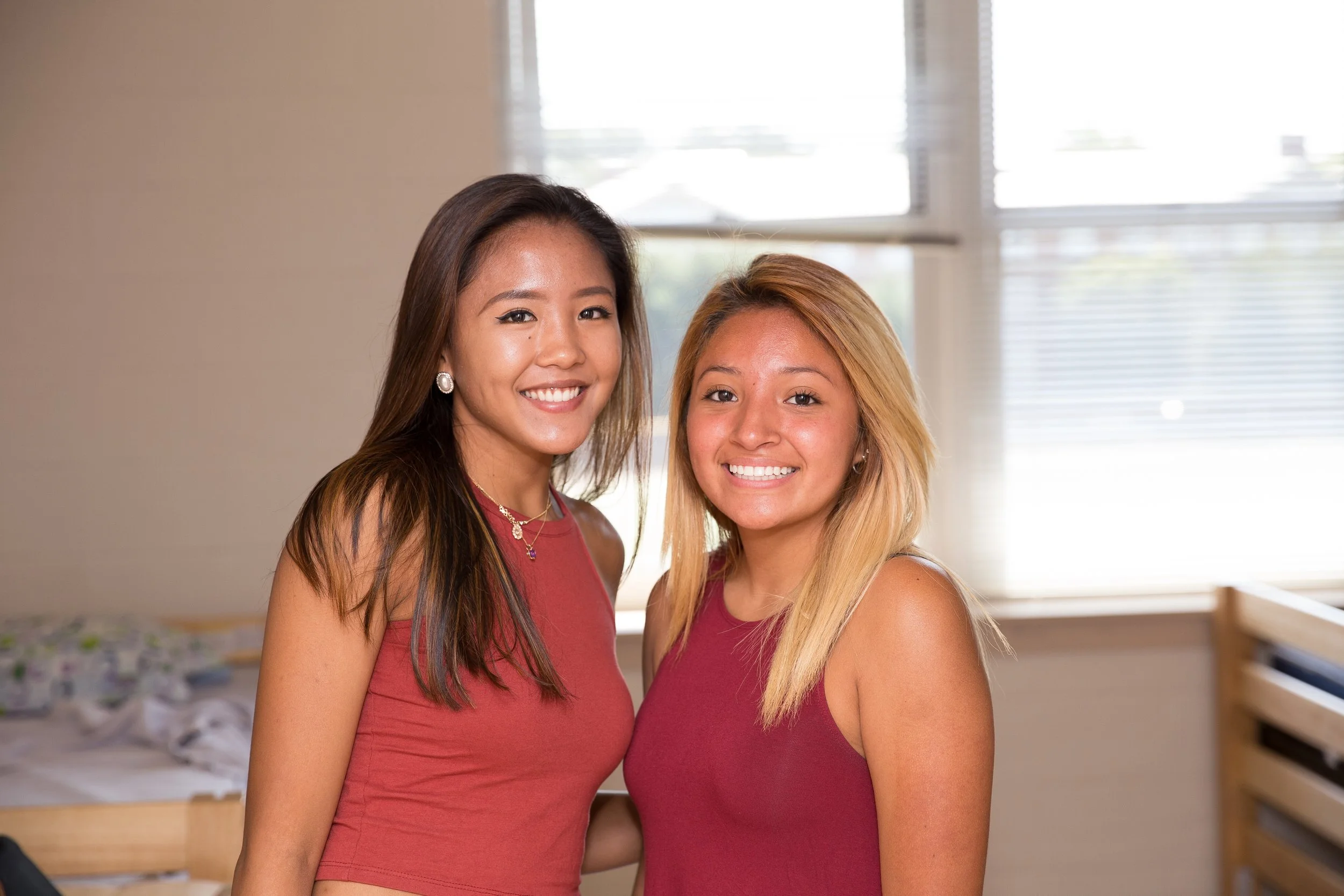 Two smiling women standing close together indoors, both wearing sleeveless maroon tops, with beds and a window with blinds in the background.