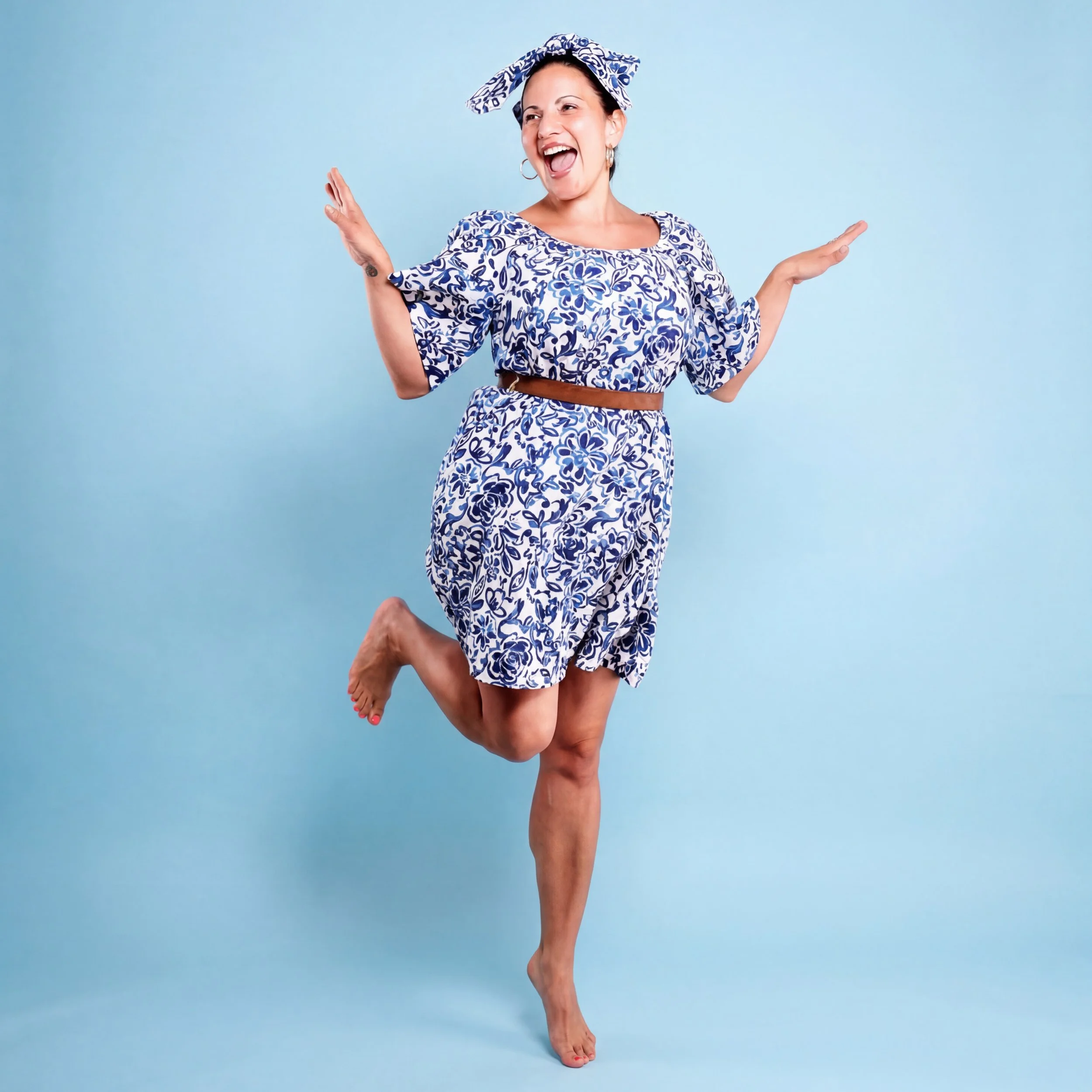 Woman smiling joyfully, wearing a matching blue and white floral dress and headscarf, standing against a light blue background.