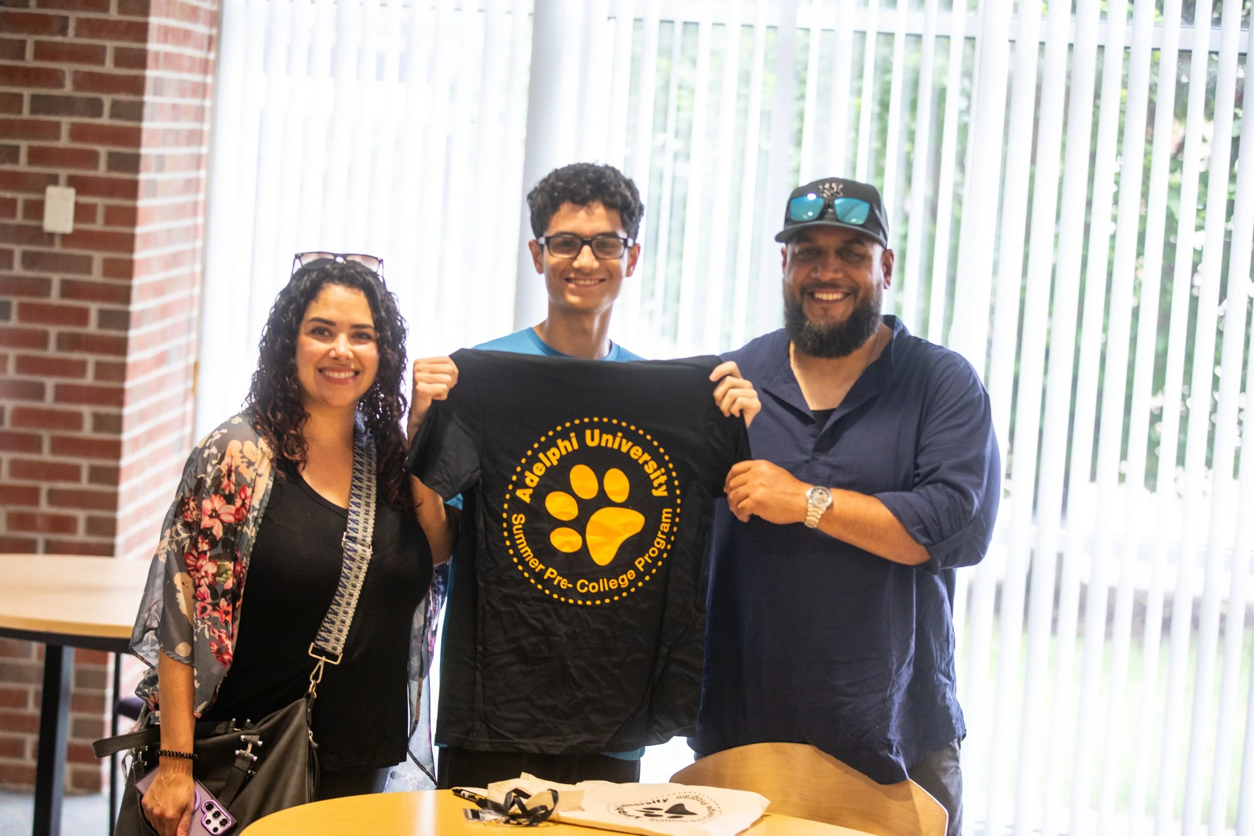Three people smiling and holding up a black t-shirt with yellow text and a paw print, inside a room with large vertical window blinds and brick wall in the background.