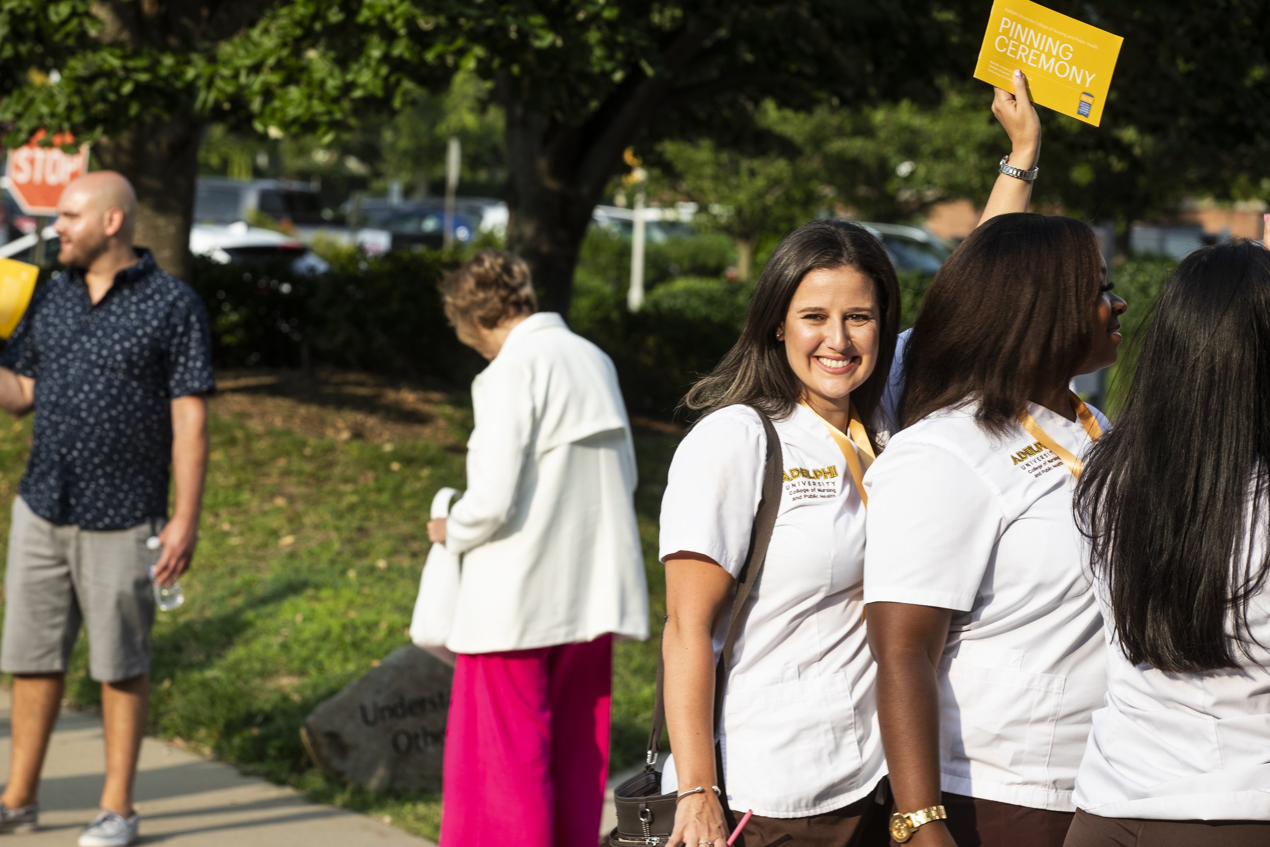 Group of people at an outdoor ceremony, some wearing white shirts with university logos, one woman smiling holding a yellow card that reads 'Pinning Ceremony', trees and parked cars in the background.
