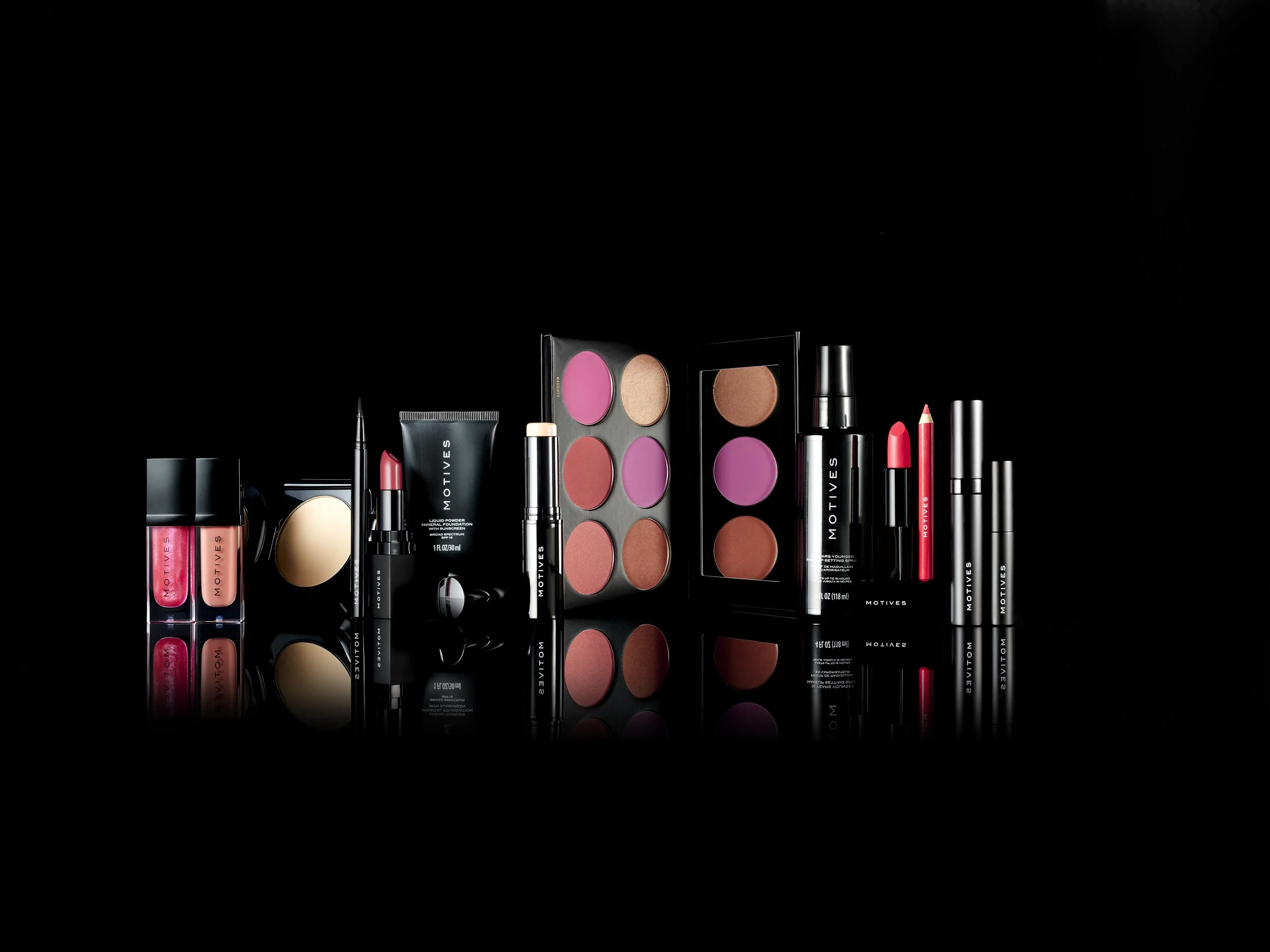 Collection of Motives cosmetics products including lip gloss, lipstick, blush, contour palette, and mascara, arranged on a reflective black surface.
