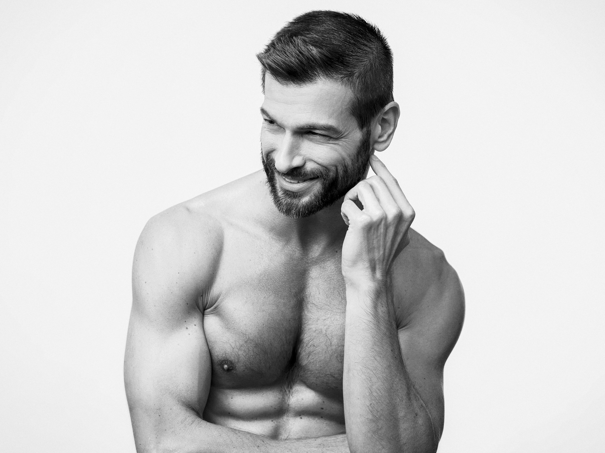 Black and white photo of a shirtless man with a beard, smiling and touching his neck, looking to his right.