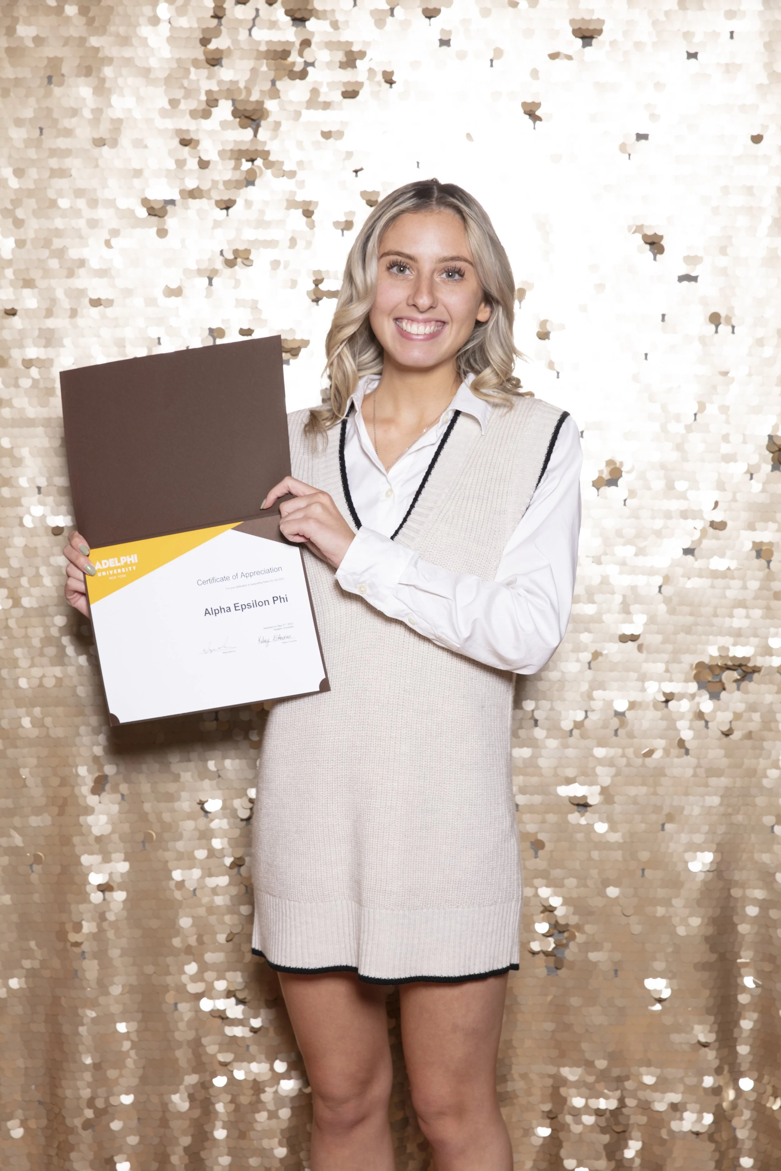 A young woman with blonde hair smiling and holding a diploma in front of a gold sequin backdrop.