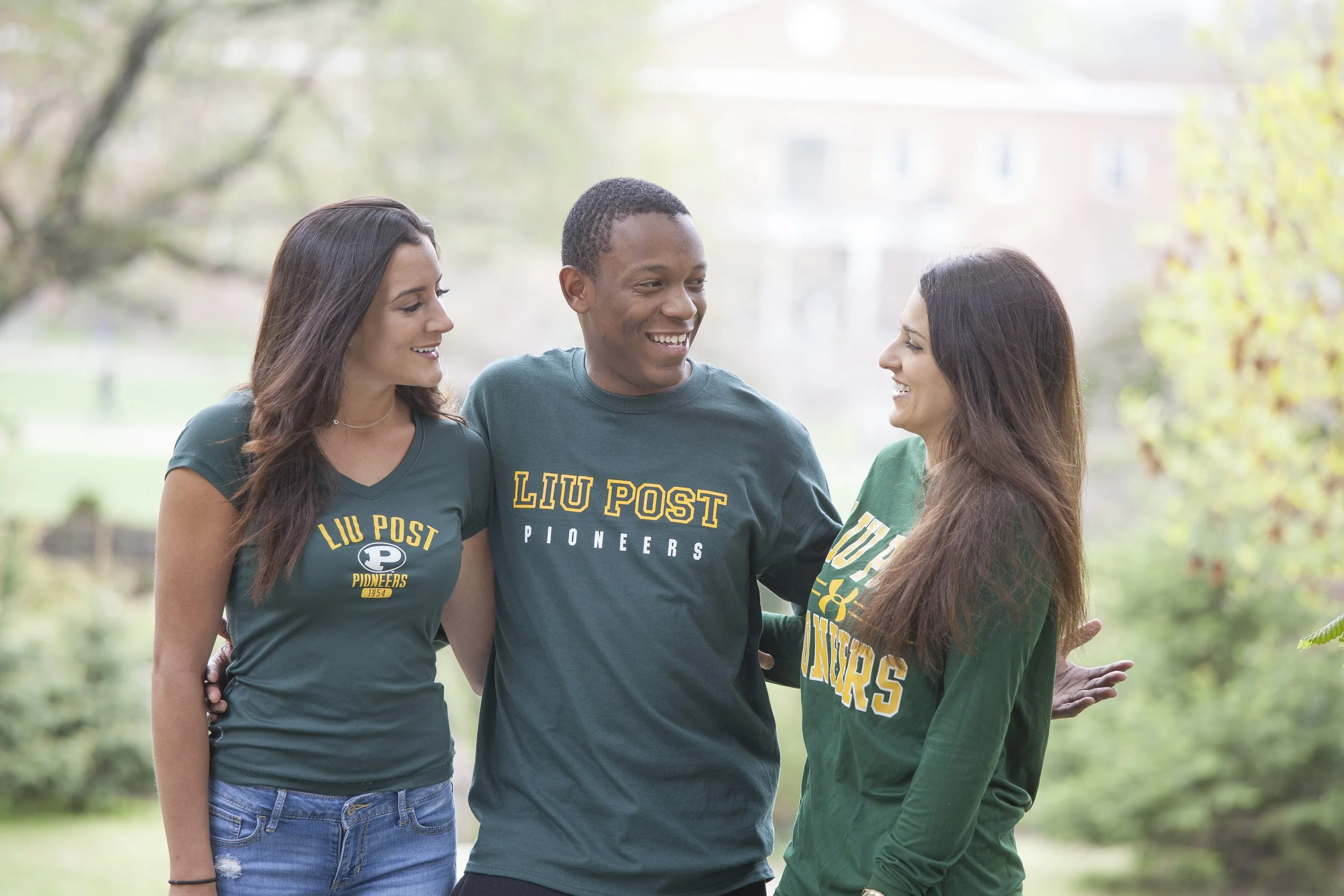 Three young adults, two women and one man, outdoors in a park, smiling and talking. They are wearing green shirts with 'Liu Post Pioneers' printed on them.