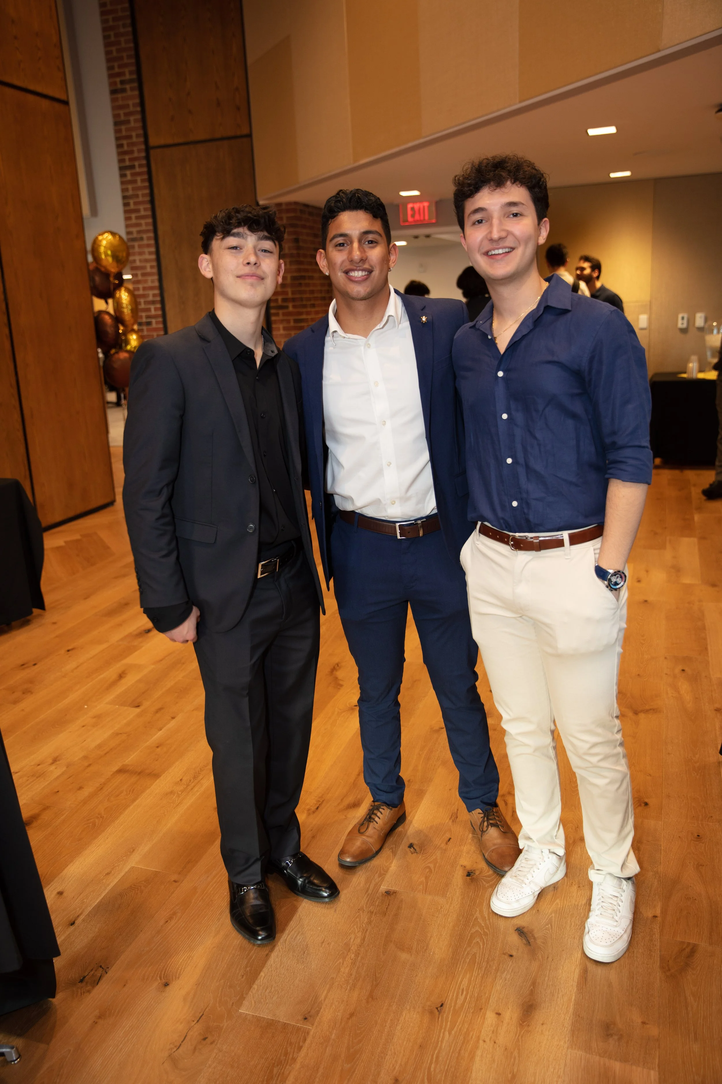 Three young men dressed in semi-formal attire standing together and smiling in an indoor event space.