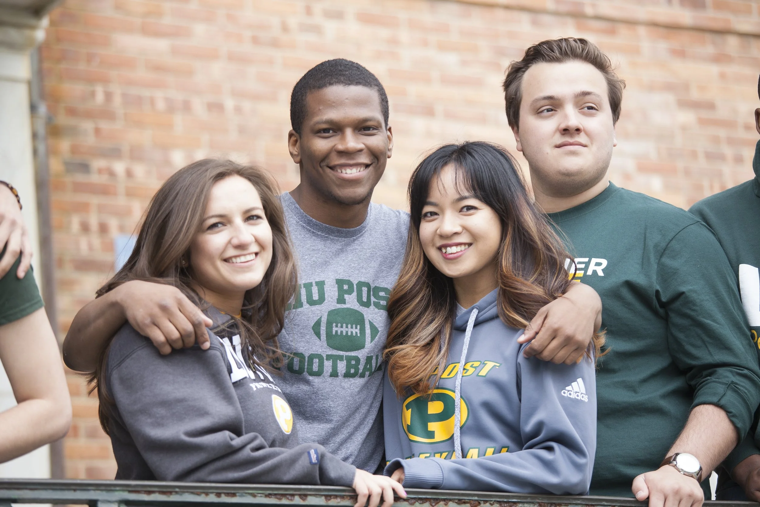 A group of four diverse young adults smiling and standing close together outdoors in front of a brick wall, wearing casual clothing and sports-related hoodies and t-shirts.