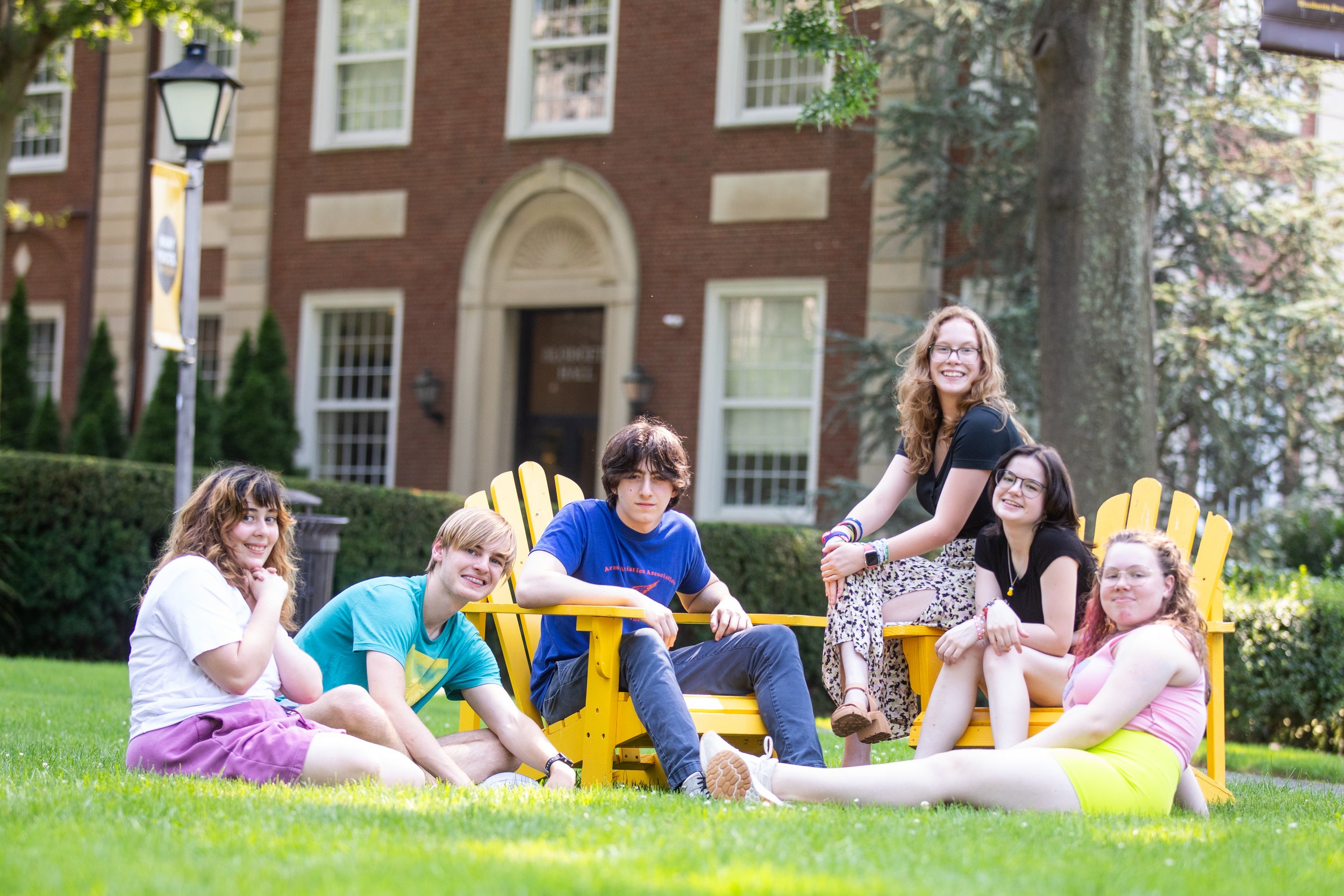 Group of six teenagers spending time outdoors on a grassy lawn with yellow chairs and a brick building in the background. Four are sitting on the grass, two are seated on chairs, and one is kneeling on the grass. They are smiling and enjoying the sun