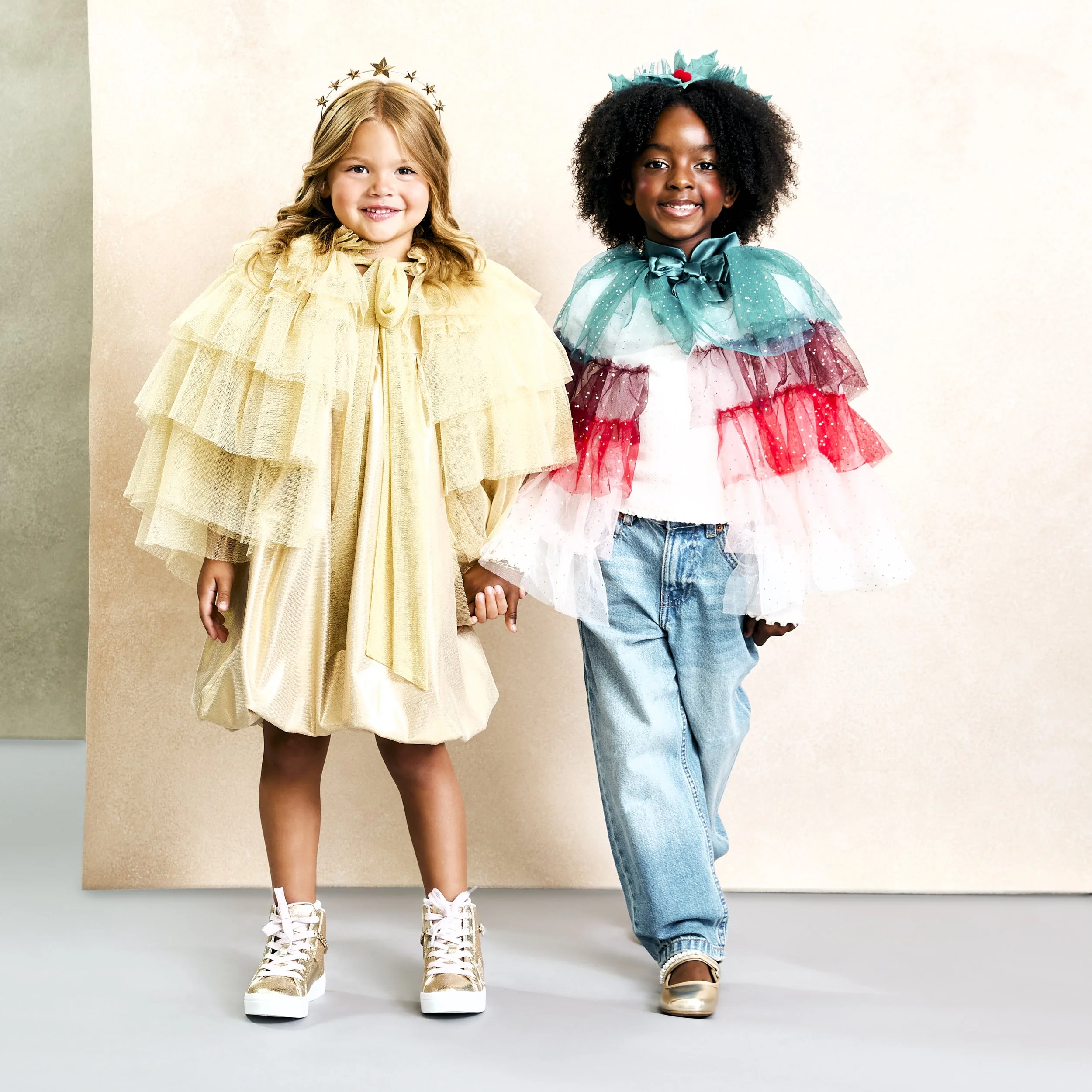 Two young girls holding hands, dressed in colorful tulle and ruffle outfits, standing on a neutral background.