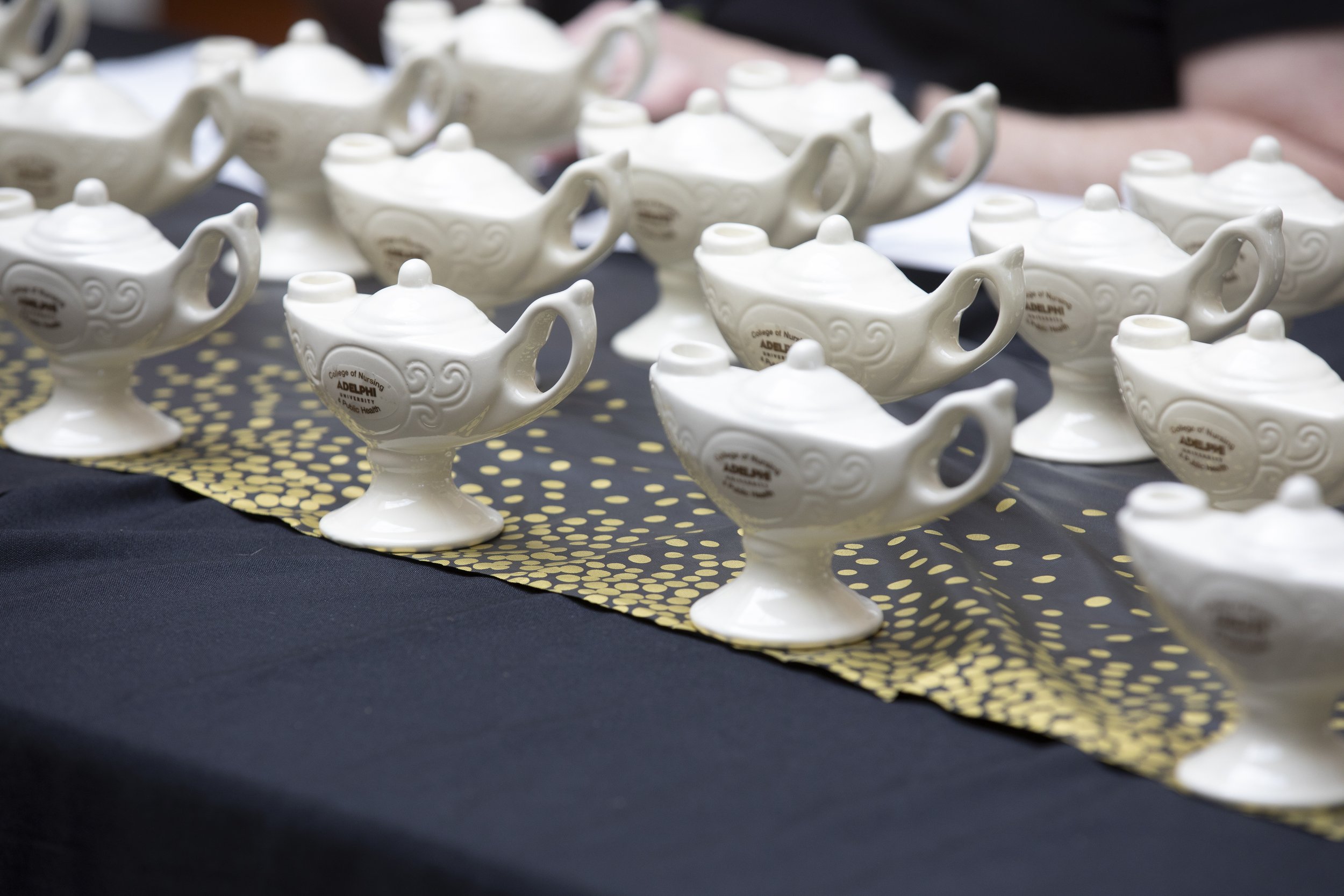Multiple white ceramic teapot-shaped awards on a black table with a gold and black patterned table runner, at an event. Some of the awards have labels from Adelphi College of Nursing.