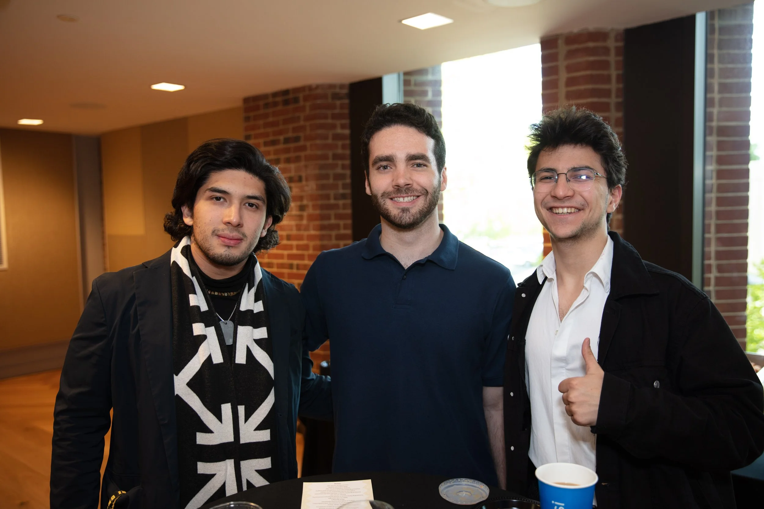 Three young men standing together indoors, smiling at the camera. The man on the left has dark hair, a scarf, and a black jacket. The man in the middle has dark curly hair and is wearing a navy polo shirt. The man on the right has glasses, dark hair,