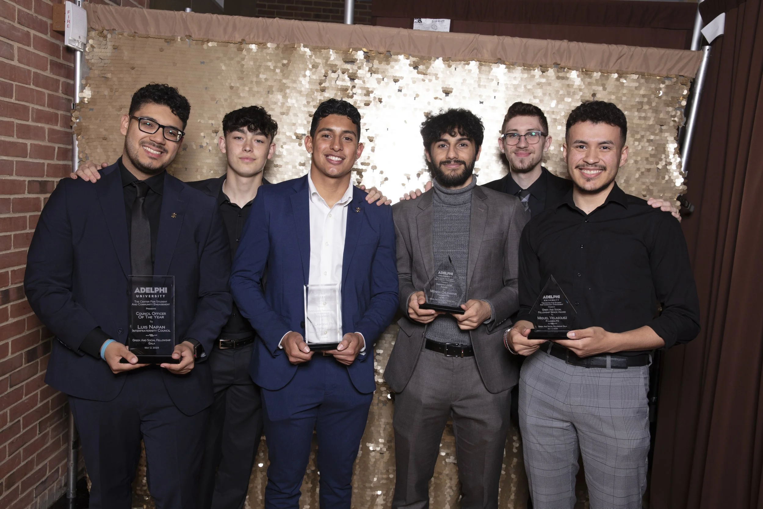 Six young men in formal attire standing together indoors, holding awards, in front of a sequin backdrop.