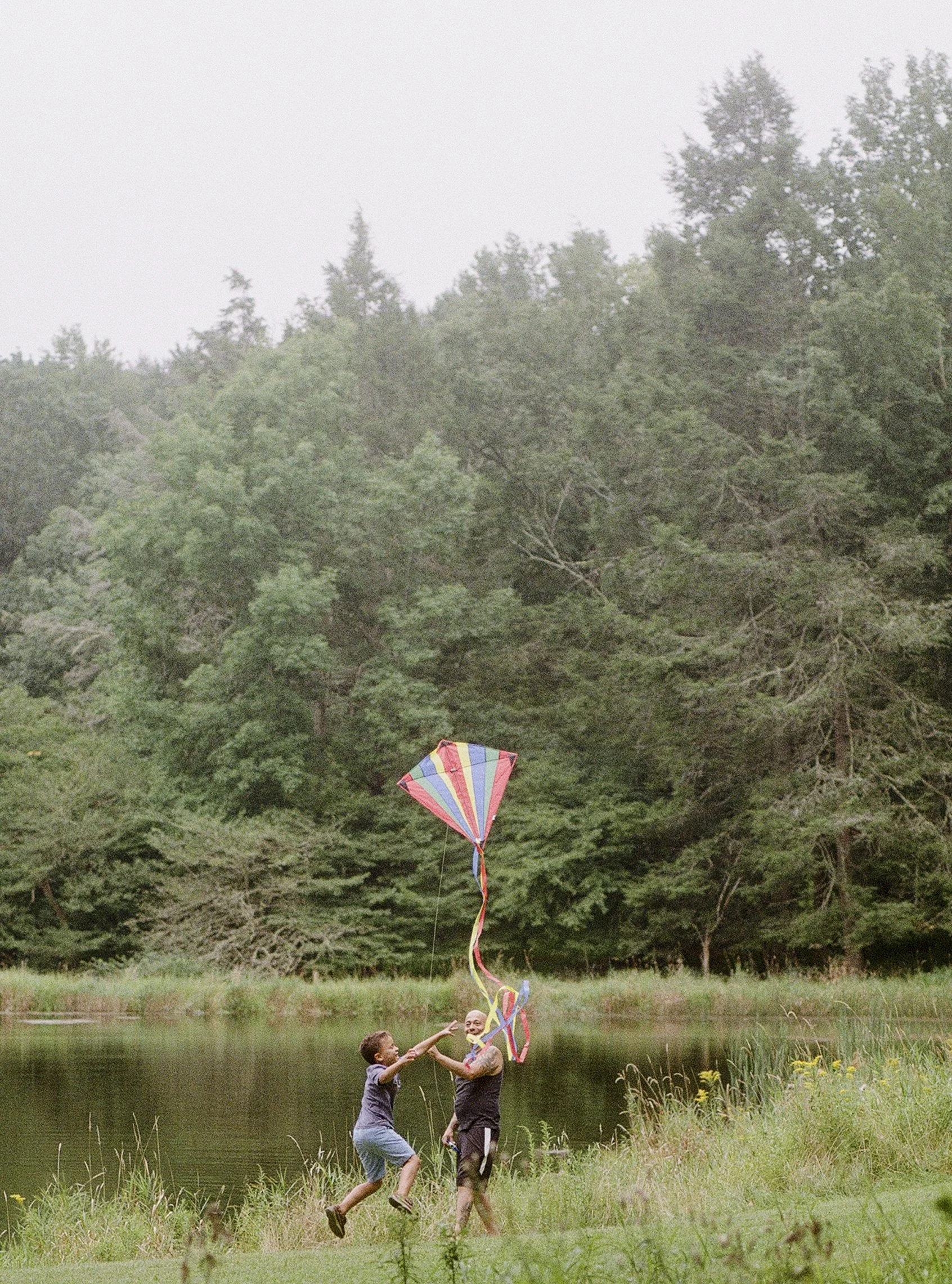 Two boys are flying a colorful kite by a lake with trees in the background. One boy is giving the kite string to the other while they are smiling and jumping.