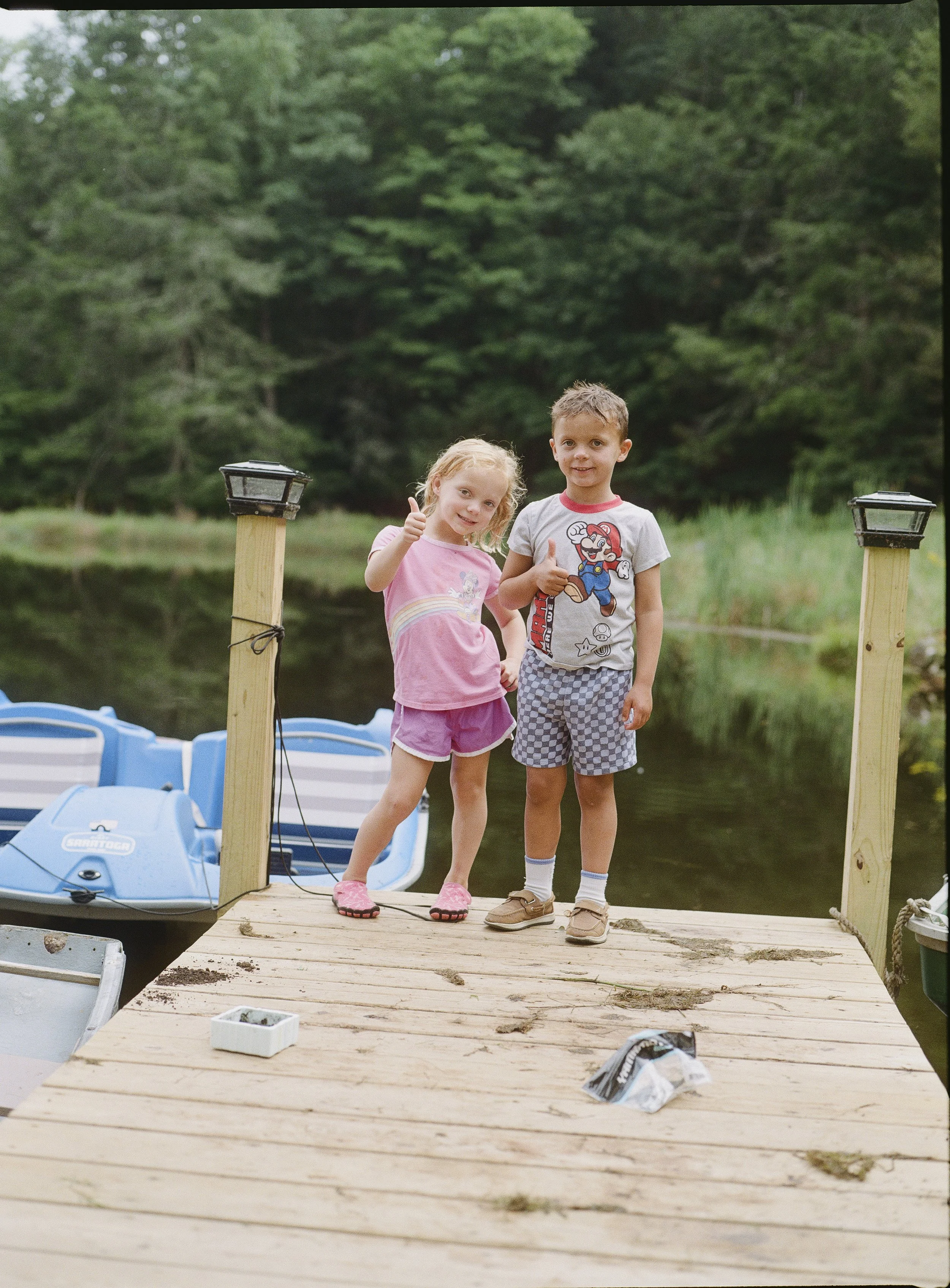 Two children, a girl and a boy, standing on a wooden dock near a body of water, with the girl giving a thumbs-up and the boy showing a thumbs-up. Behind them are two wooden posts, a blue paddle boat, and green trees in the background.