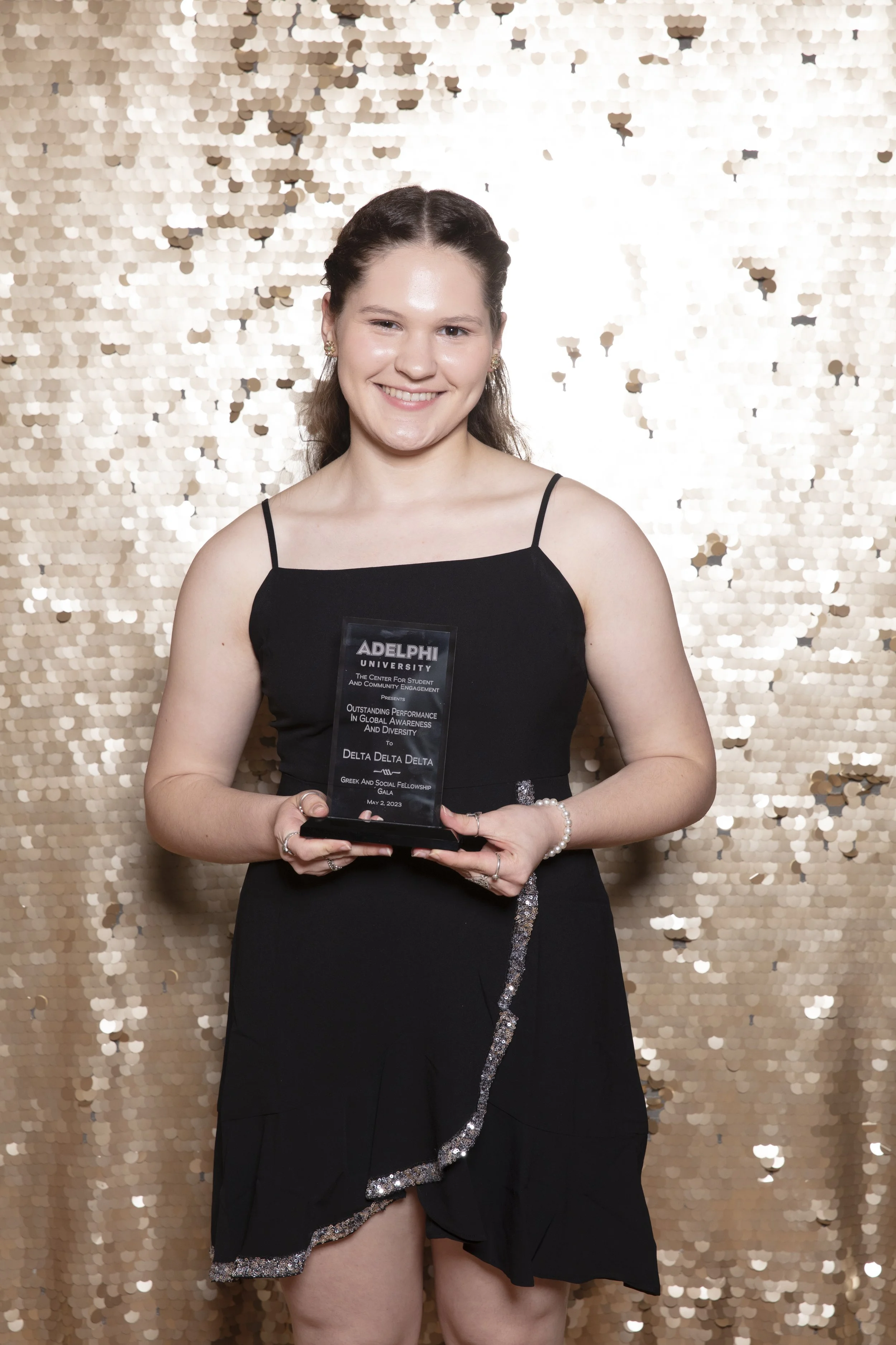 Young woman in black dress holding an award, standing in front of a gold sequin backdrop, smiling at the camera.
