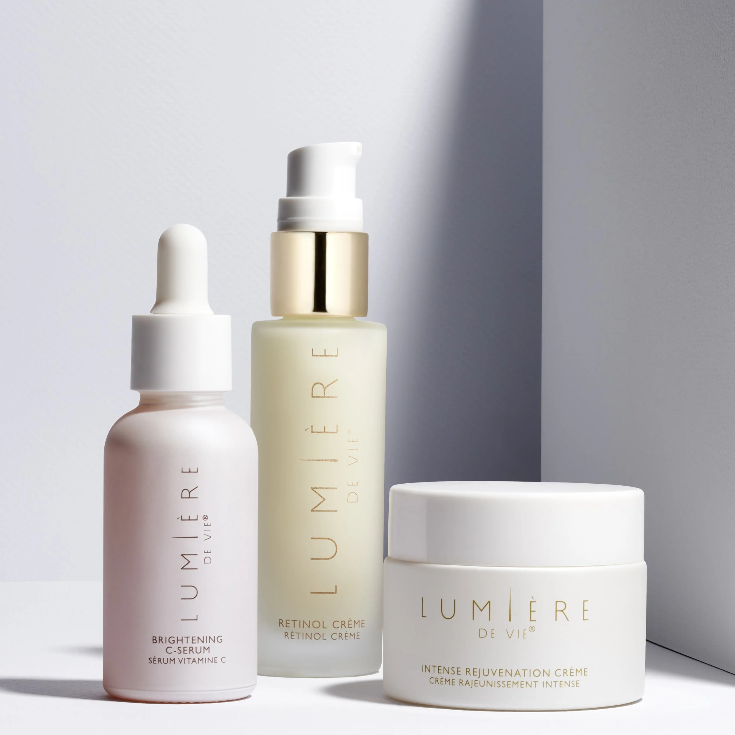 Three skincare products from Lumière de Vie: a Brightening C-Serum in a pink bottle, a Retinol Crème in a clear bottle, and an Intense Rejuvenation Crème in a white jar, arranged on a white surface with a minimalist background.