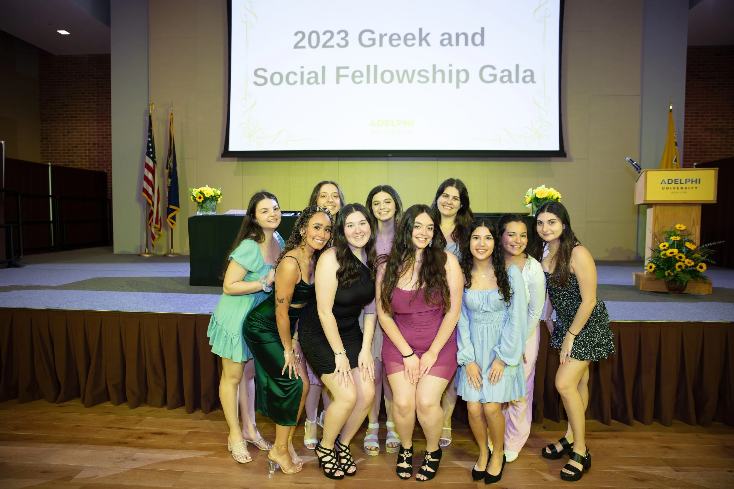 Group of young women in dresses posing together on a stage at the 2023 Greek and Social Fellowship Gala at Adelphi University, with a large screen and flags in the background.