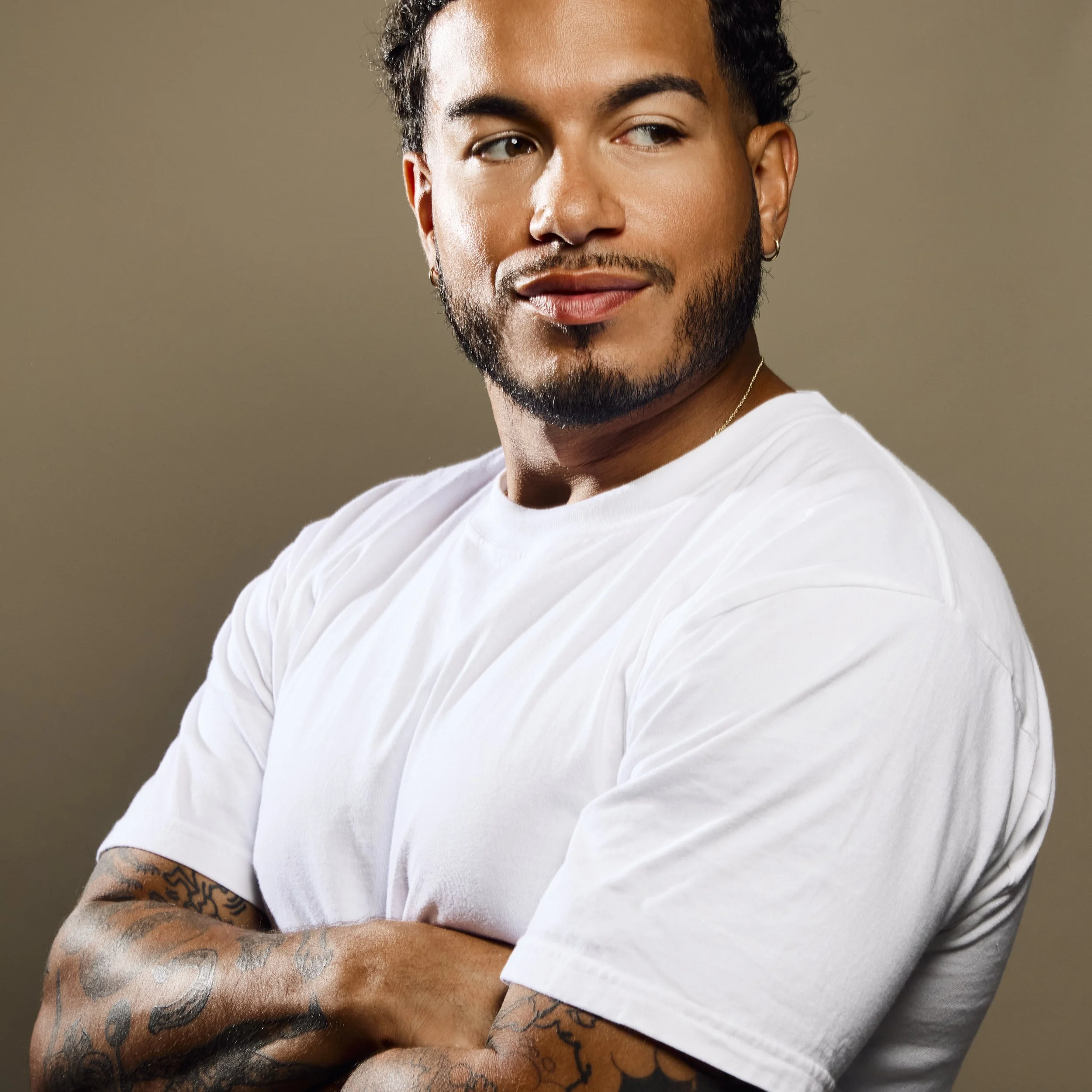 Close-up of a young man with dark curly hair, tattoos on his arms, earrings, and a white T-shirt, looking to the side against a plain background.