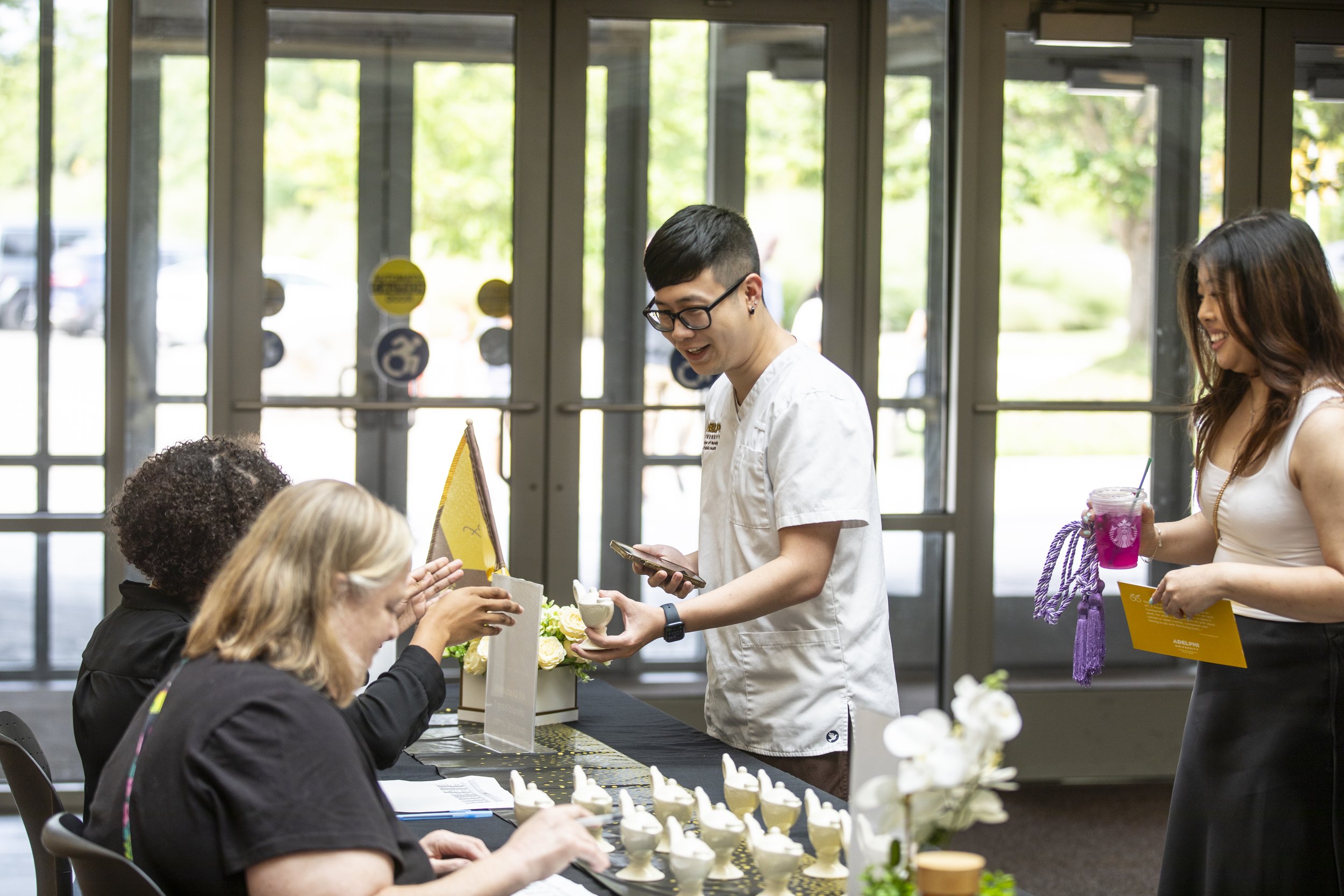 People at a registration table for a workshop or event, with a woman in a white dress holding a pink drink, a young man in a white chef coat exchanging a small bowl, and two women seated, one with blonde hair and one with curly hair, engaging with th