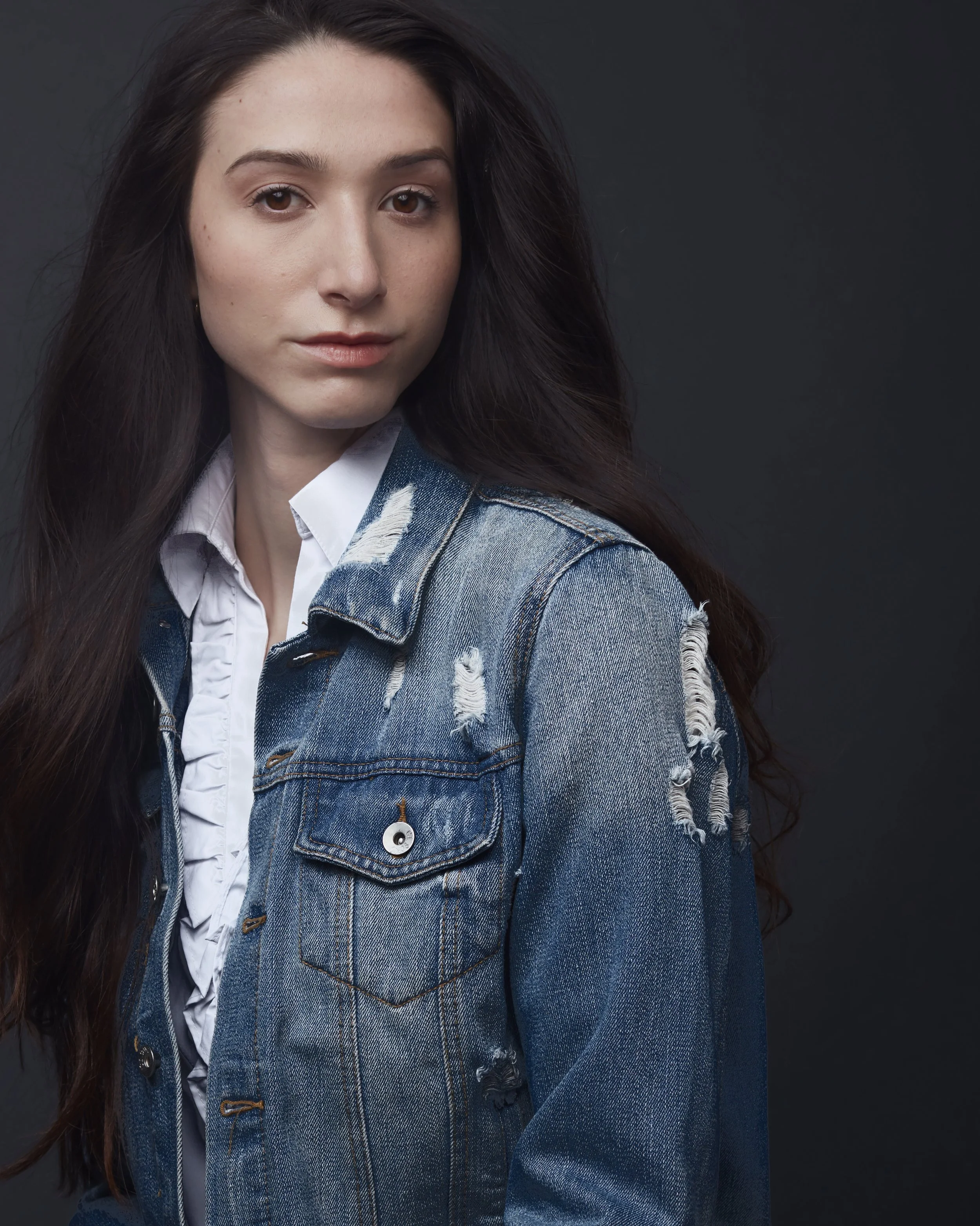 A young woman with long dark hair wearing a white shirt and a distressed denim jacket against a dark background.