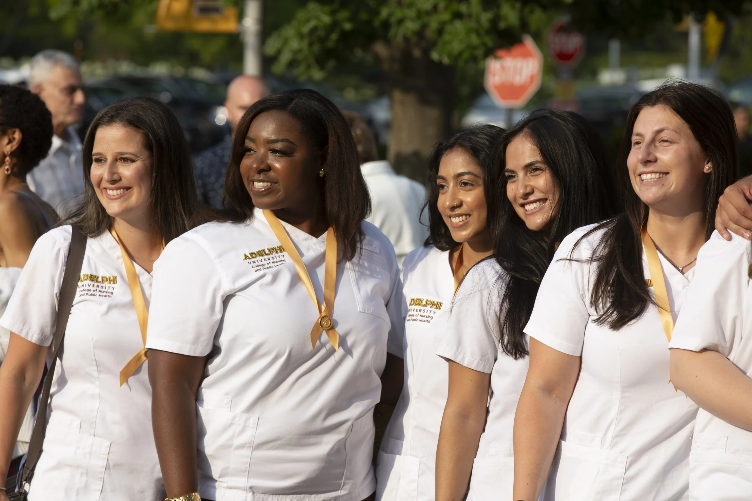 Group of diverse women in white uniforms with yellow lanyards standing outside smiling at an event.
