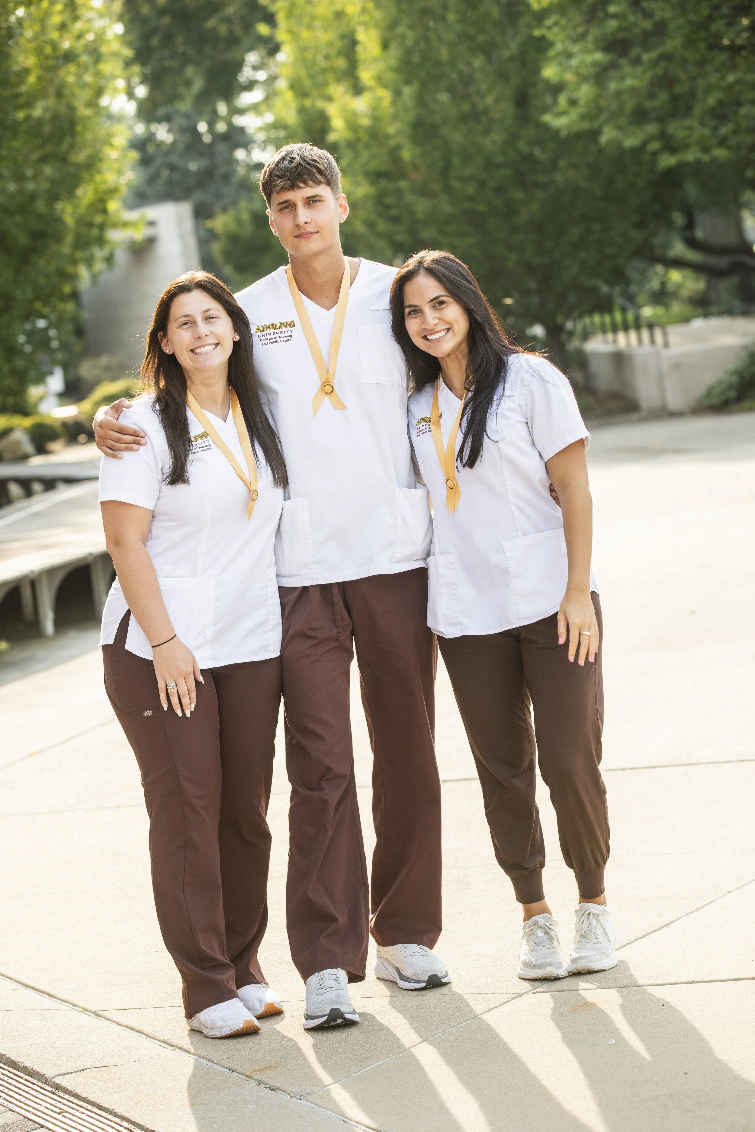 Three healthcare students wearing white coats and medals, standing outdoors on a paved path with greenery in the background.