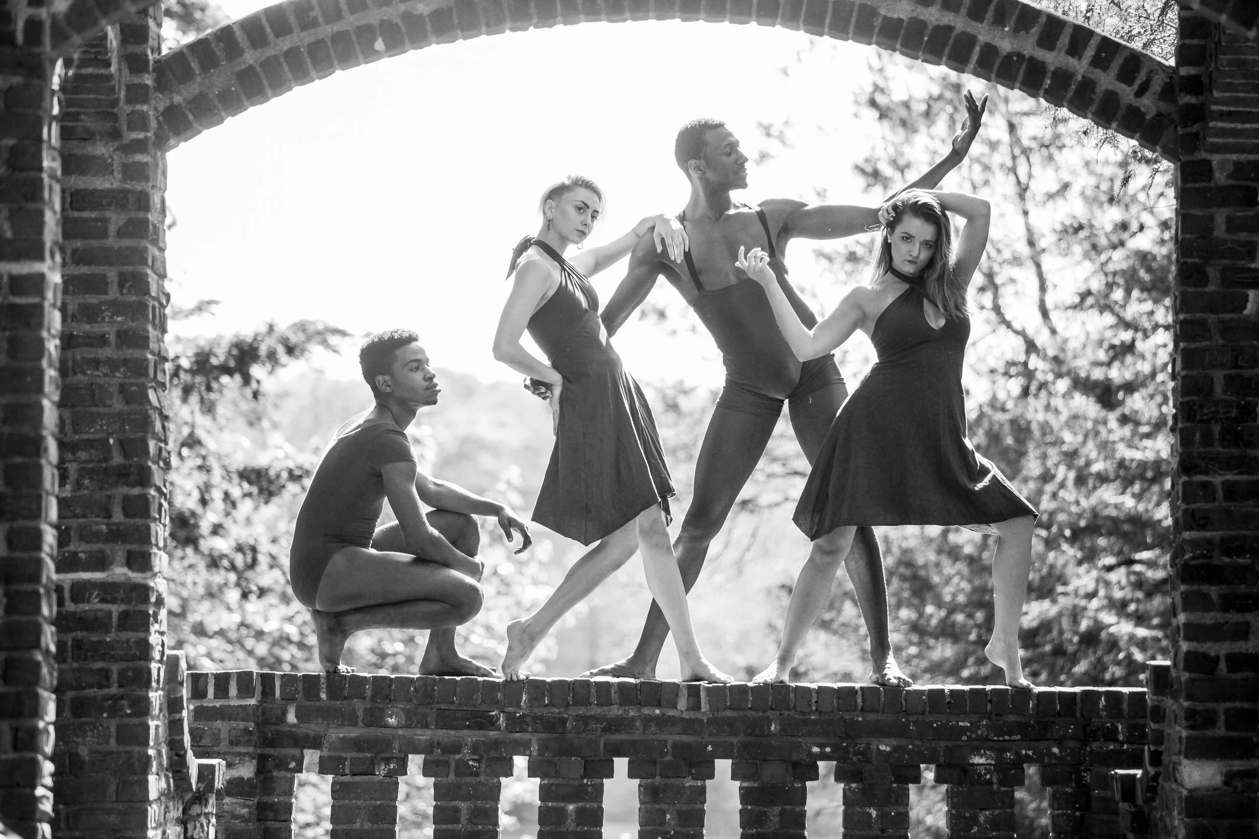 Group of five dancers, four women and one man, posing on a brick structure outdoors, with trees in the background, captured in black and white.