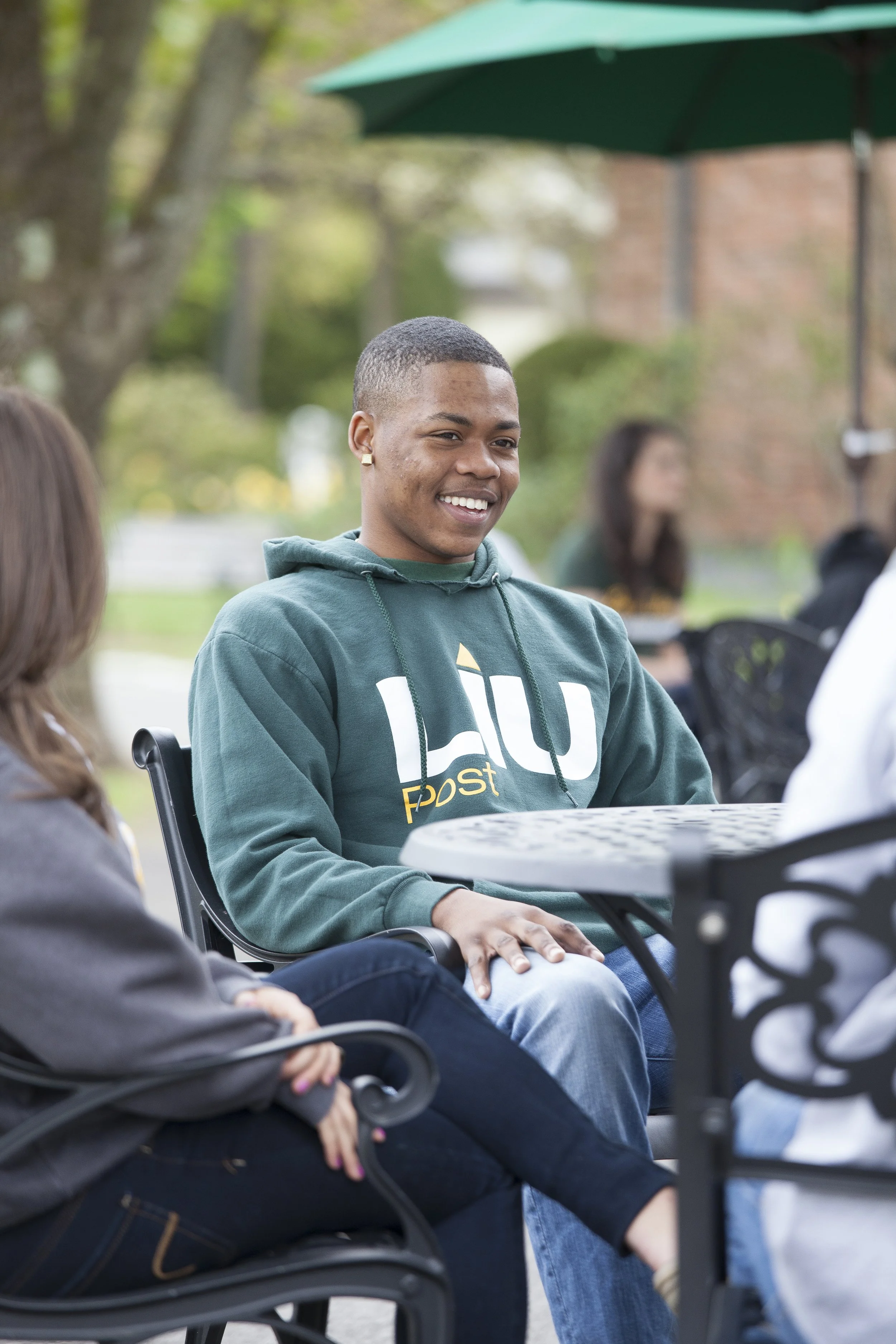 Young man wearing a green hoodie smiling and talking with a woman outdoors at a table.