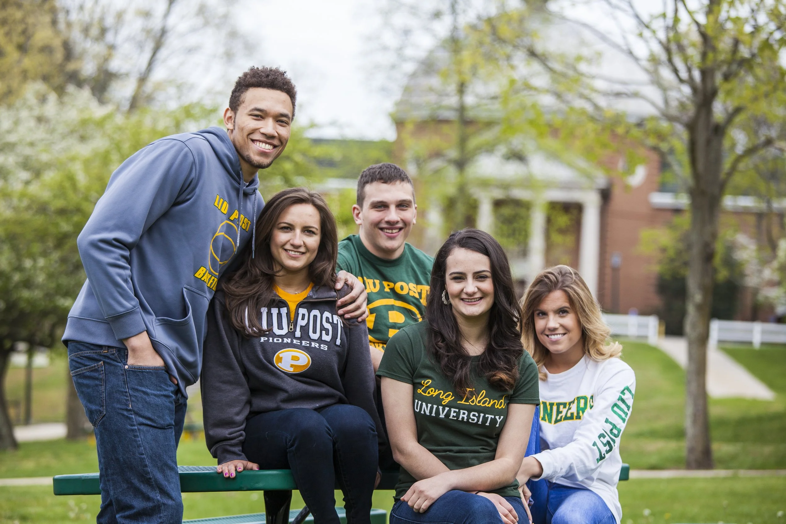 Group of five young adults sitting and standing on a park bench, smiling, with trees and a brick building in the background.